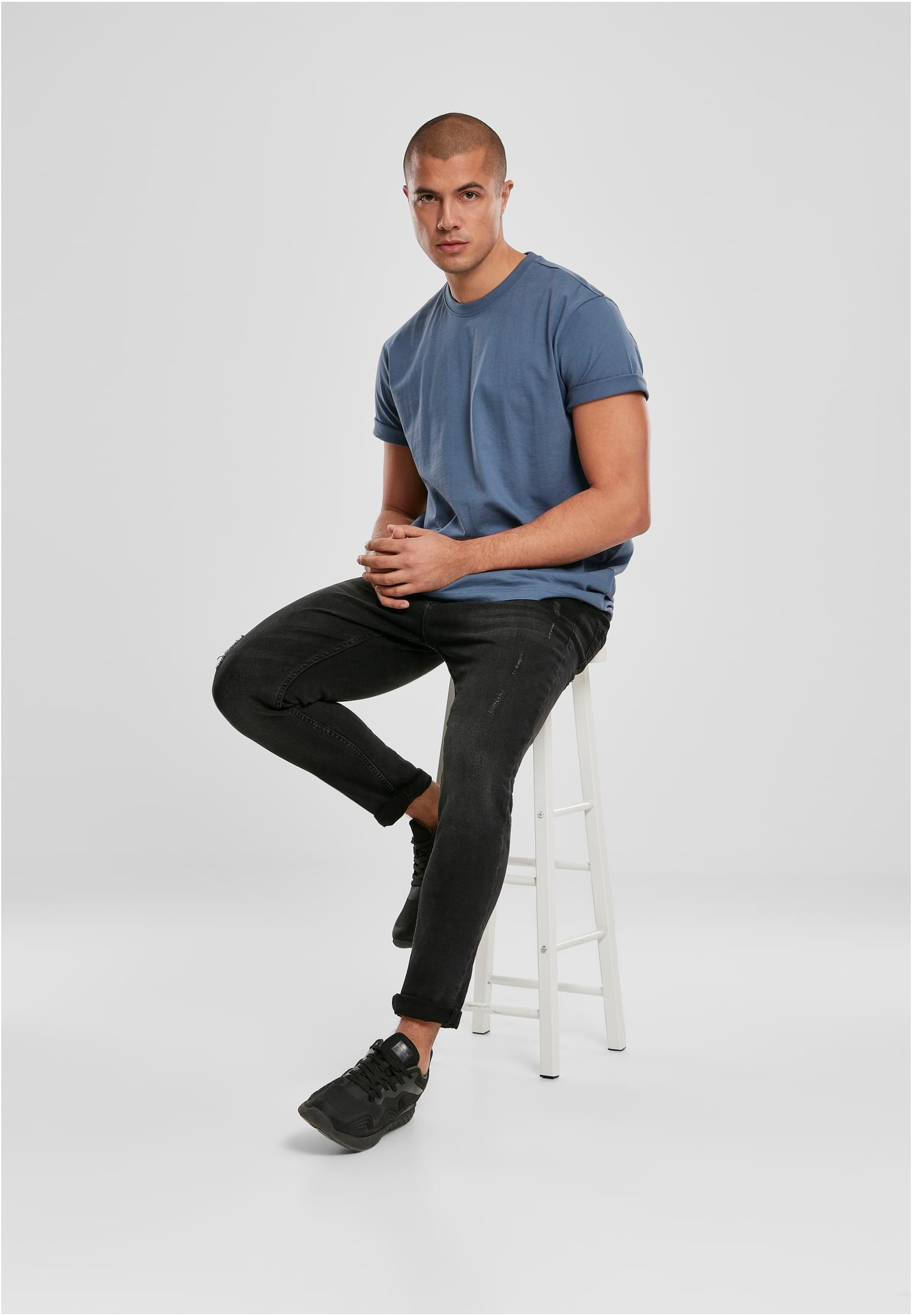 En man som bär en blå t-shirt och svarta jeans sitter på en vit pall.