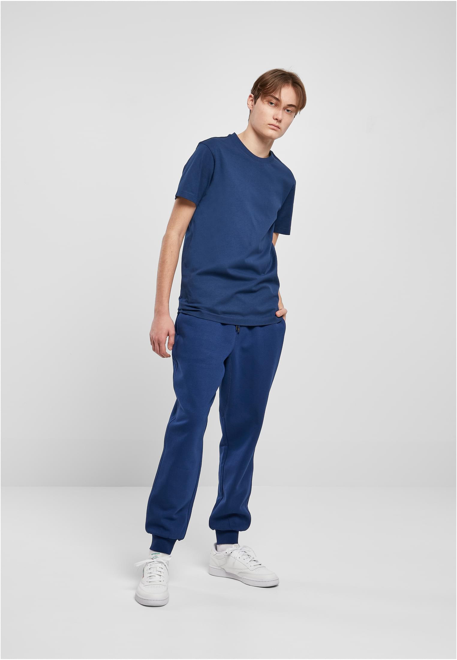 En manlig modell poserar i en blå t-shirt, matchande blå mjukisbyxor och vita sneakers mot en ljusgrå bakgrund.