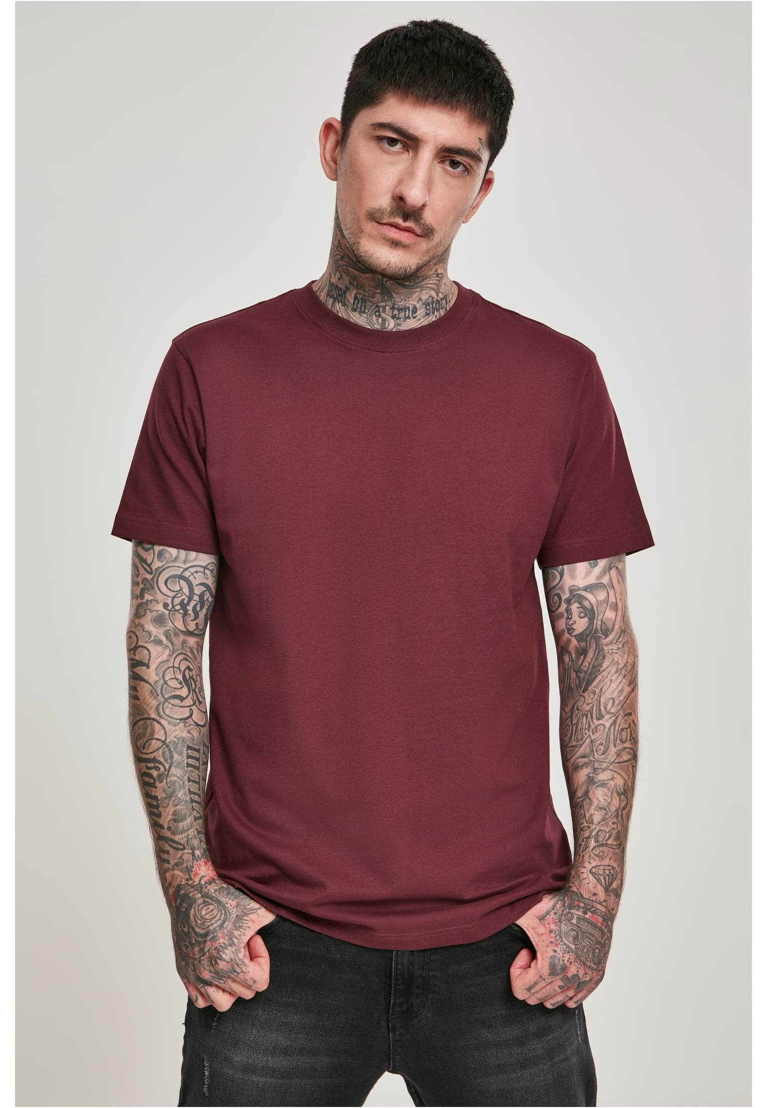 En man med tatueringar visar upp framsidan av en vinröd t-shirt.
