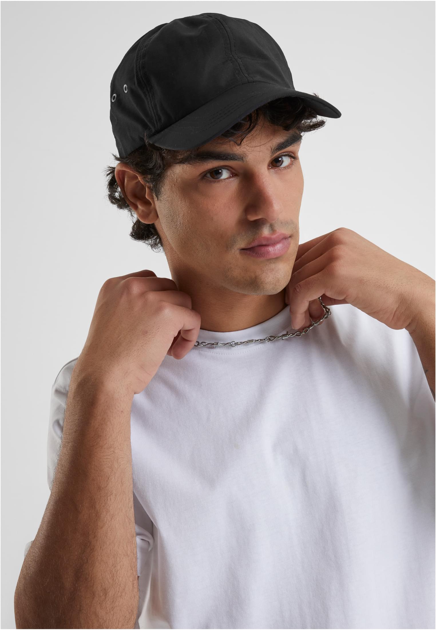 En man i vit t-shirt och svart keps justerar sitt silverhalsband medan han tittar in i kameran.