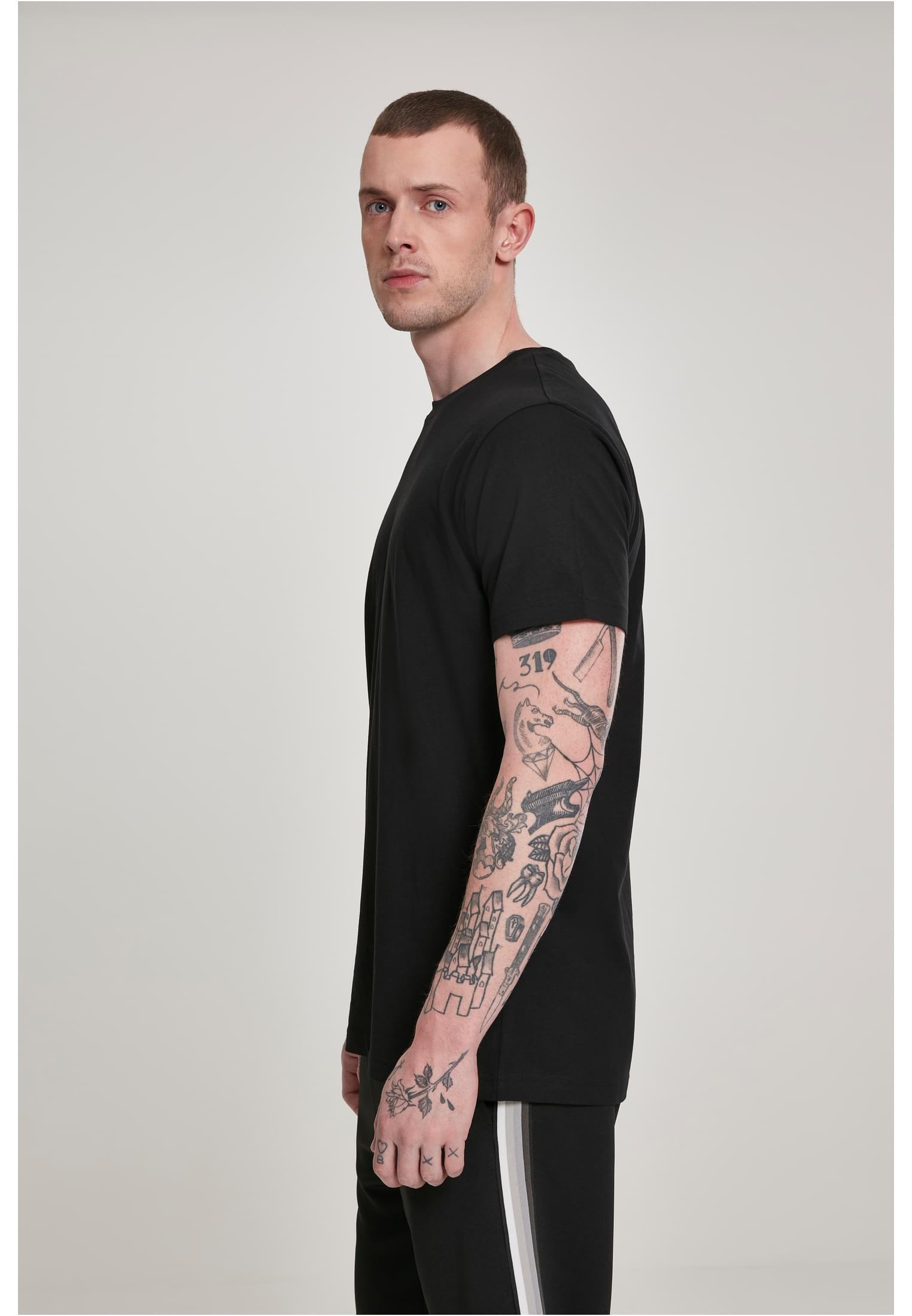 En man med en tatuerad arm poserar i en svart Basic Tee, sedd från sidan.
