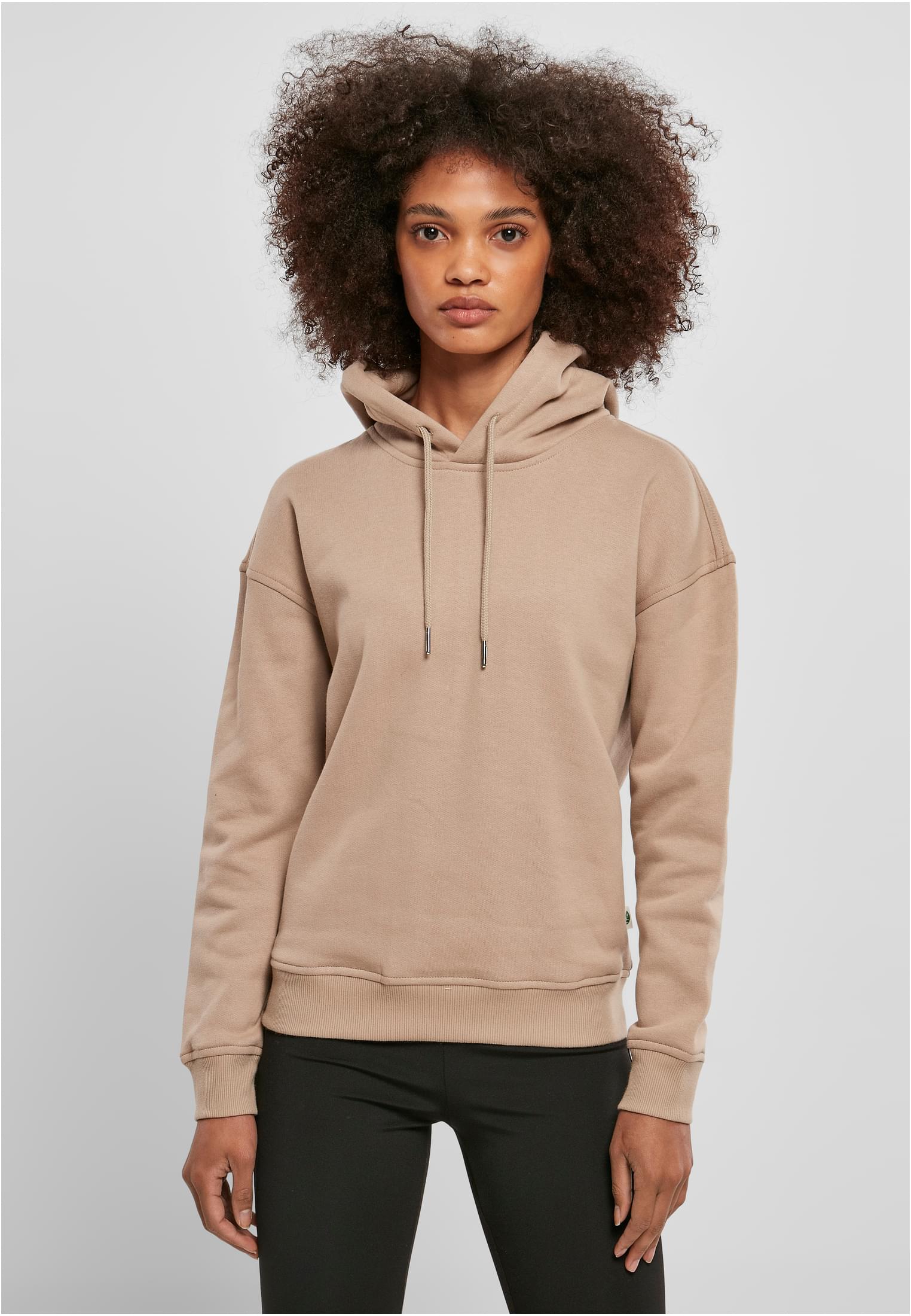 En kvinna med lockigt hår bär en beige UC Ladies Organic Hoody och tittar in i kameran mot en grå bakgrund.