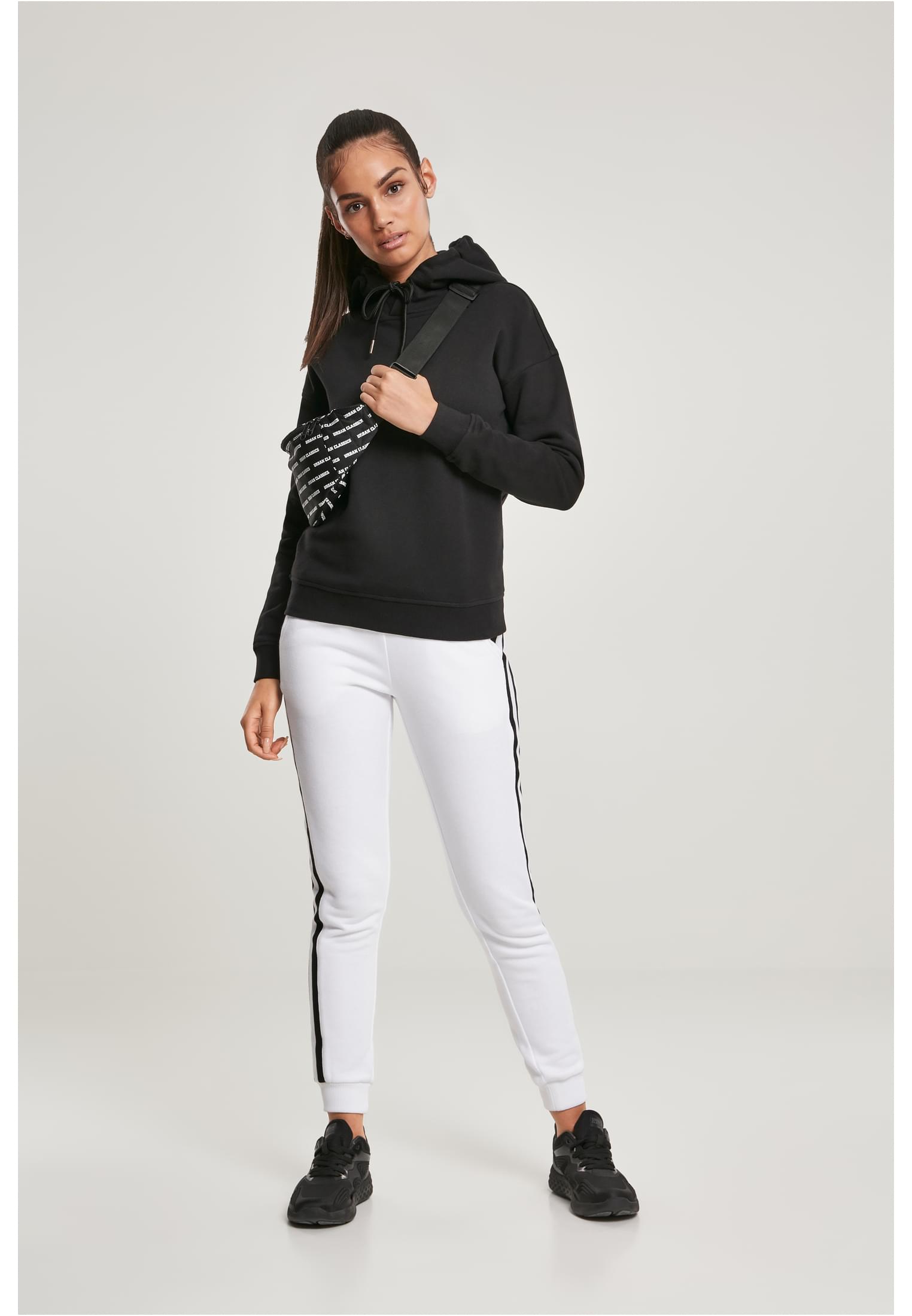 Kvinna som bär en svart UC Ladies Organic Hoody, vita mjukisbyxor med svarta ränder och svarta sneakers.