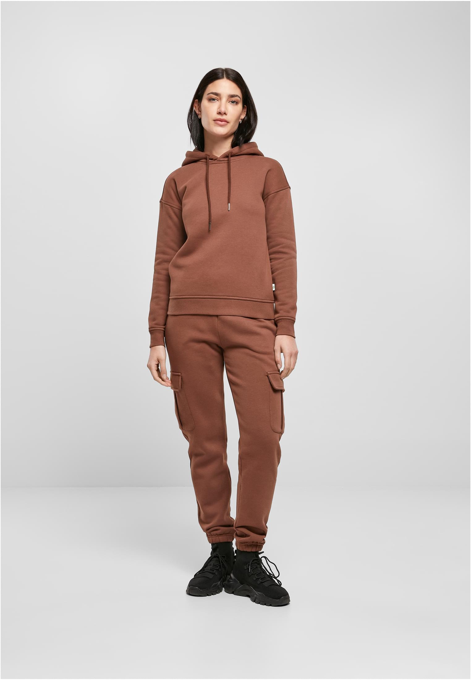 En kvinnlig modell visar framifrån UC Ladies Organic Hoody i brunt, stylad med matchande byxor, mot en ljusgrå bakgrund.