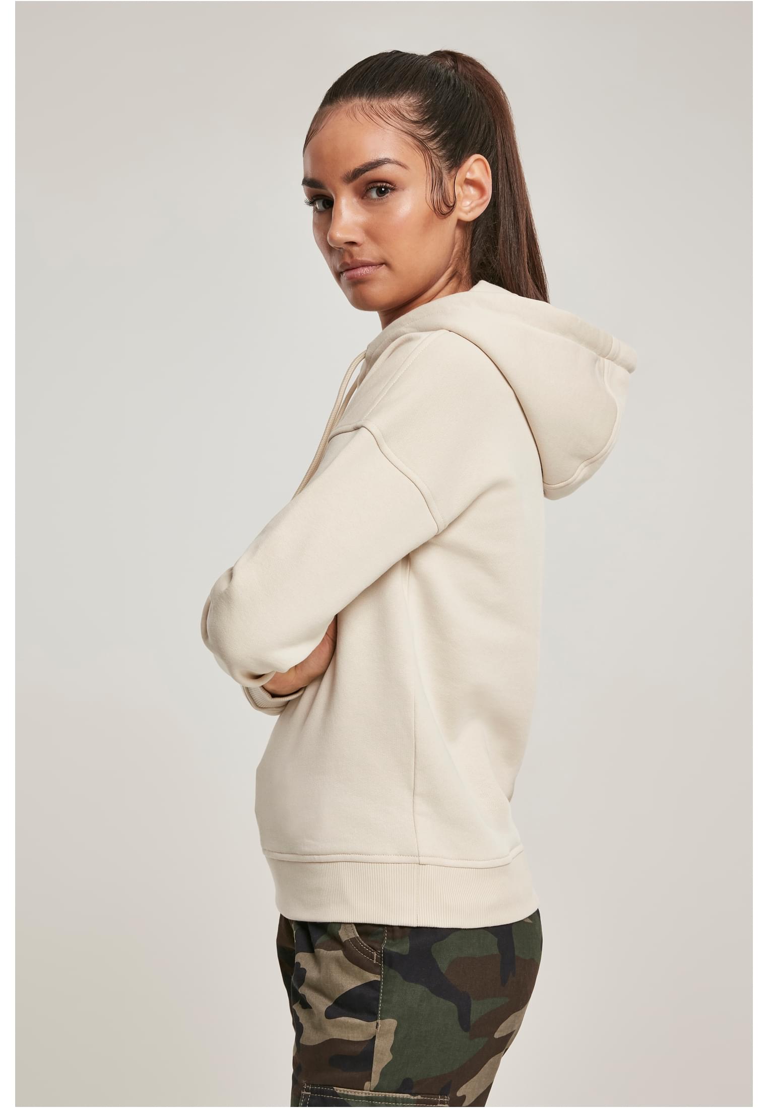 Kvinna i profil som bär en ljusbeige Ladies Organic Hoody och kamouflagebyxor. Hon har armarna i kors och tittar över axeln.