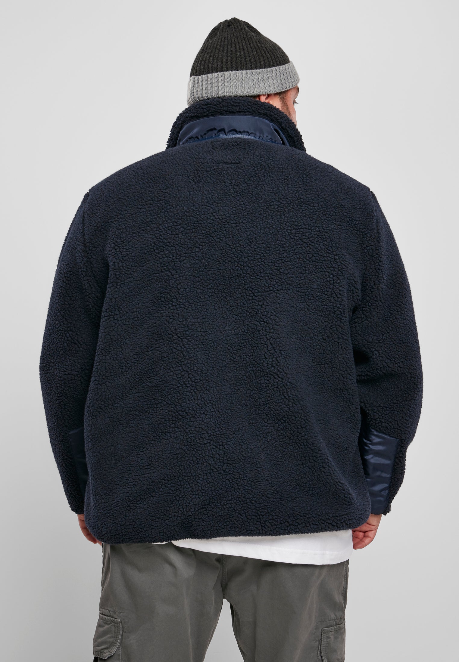 En man, sedd bakifrån, klädd i en mörkblå UC Sherpa Jacket, gråa byxor och en mörkgrå mössa.