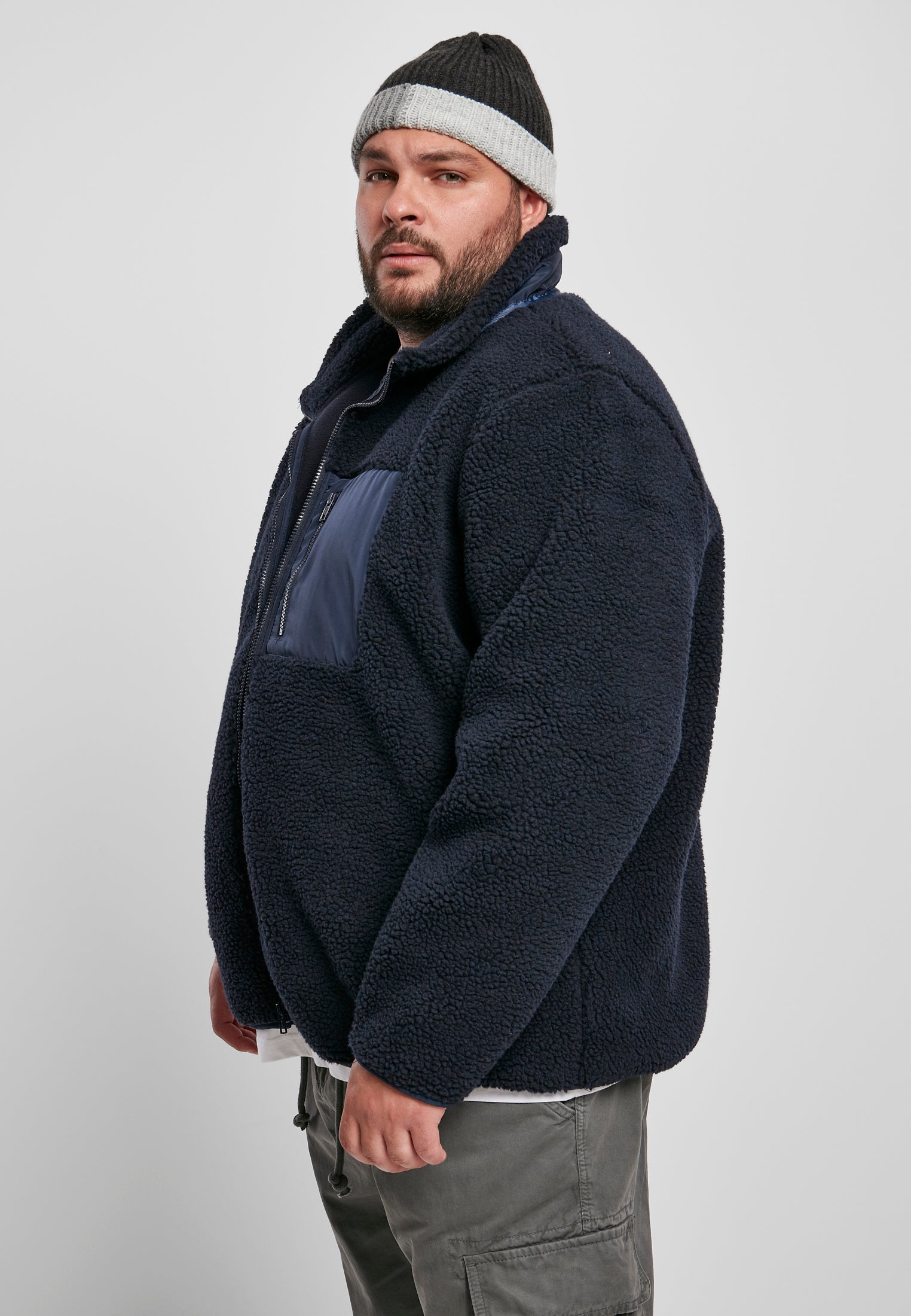 En man, sedd snett framifrån, bär en marinblå UC Sherpa Jacket med en bröstficka i nylon och en grå mössa.