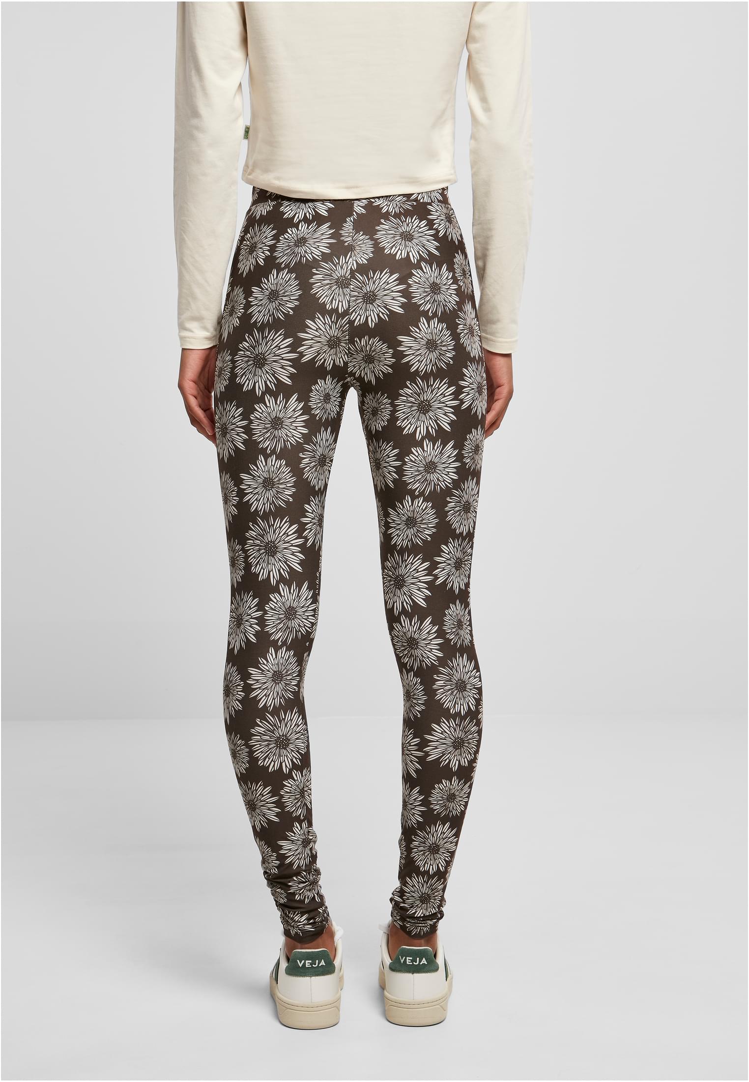Baksidan av en kvinna som bär UC Ladies Soft AOP Leggings. Leggingsen är mörkbruna med ett heltäckande mönster av vita blommor och hon bär dem med en benvit långärmad tröja och vita sneakers.