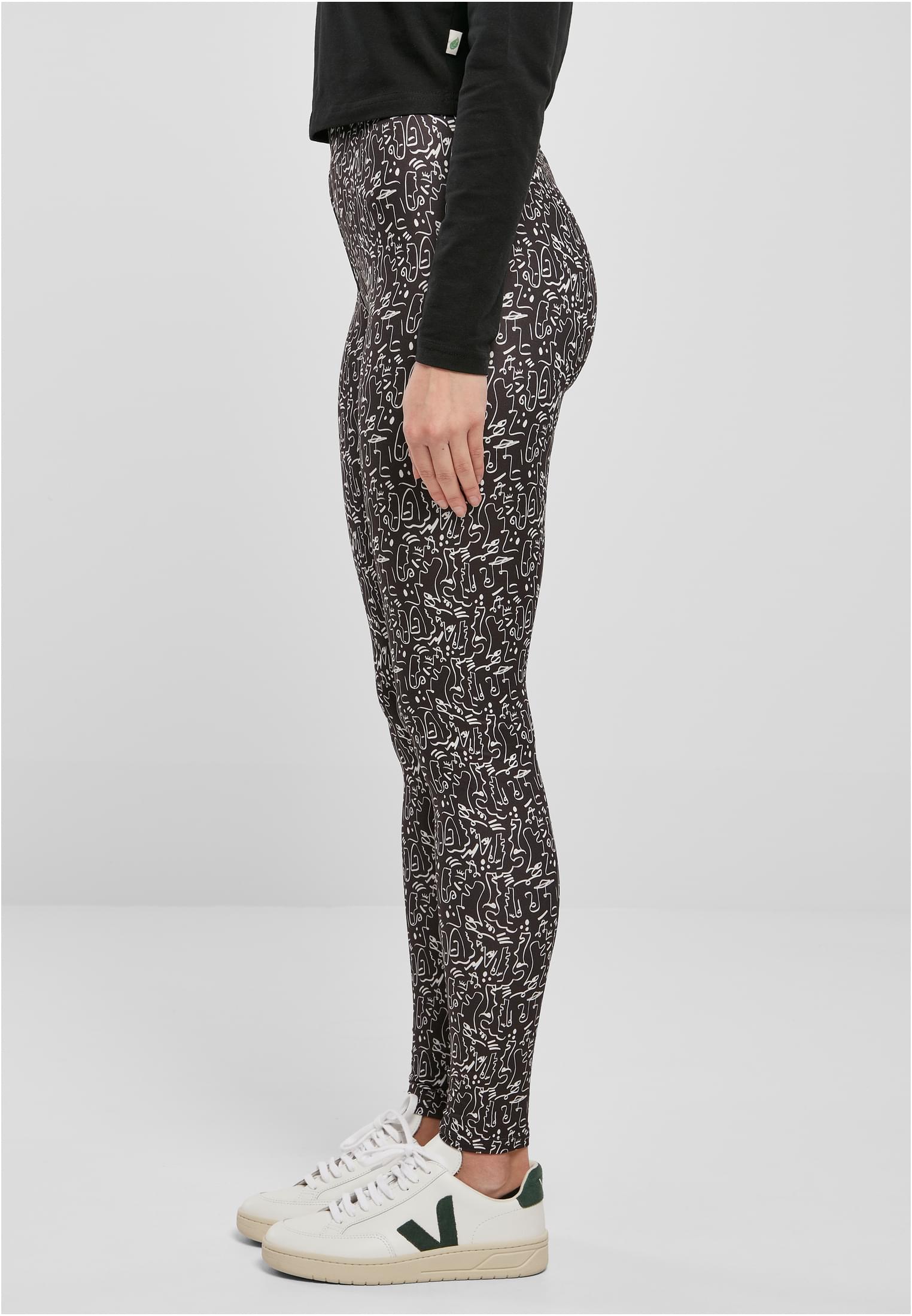 Sidovy av en kvinna som bär svarta UC Ladies Soft AOP Leggings med ett vitt heltäckande mönster, tillsammans med en svart långärmad tröja och vita sneakers.