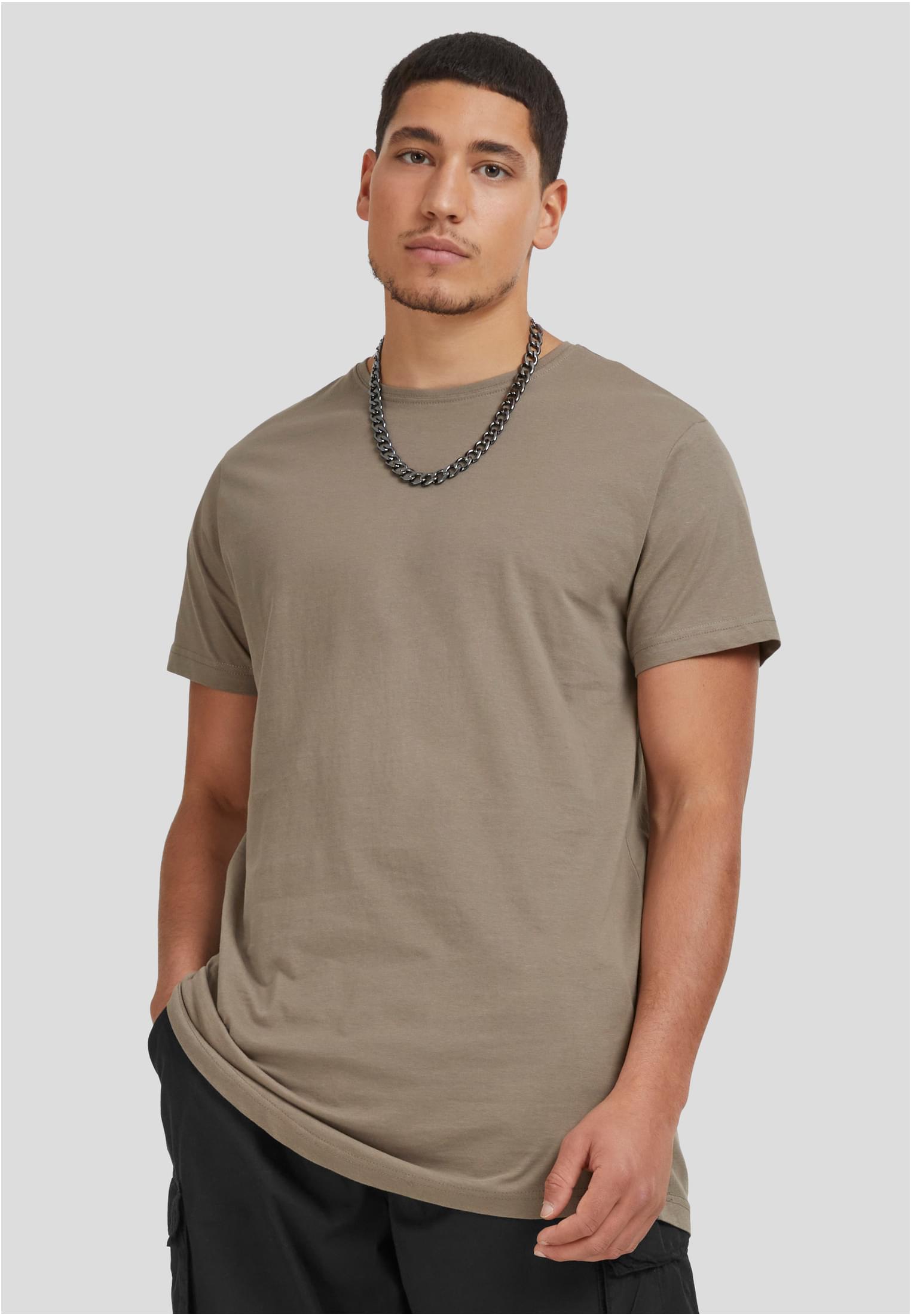 En man poserar mot en grå bakgrund, klädd i en terracottafärgad UC Shaped Long Tee och ett kedjehalsband.