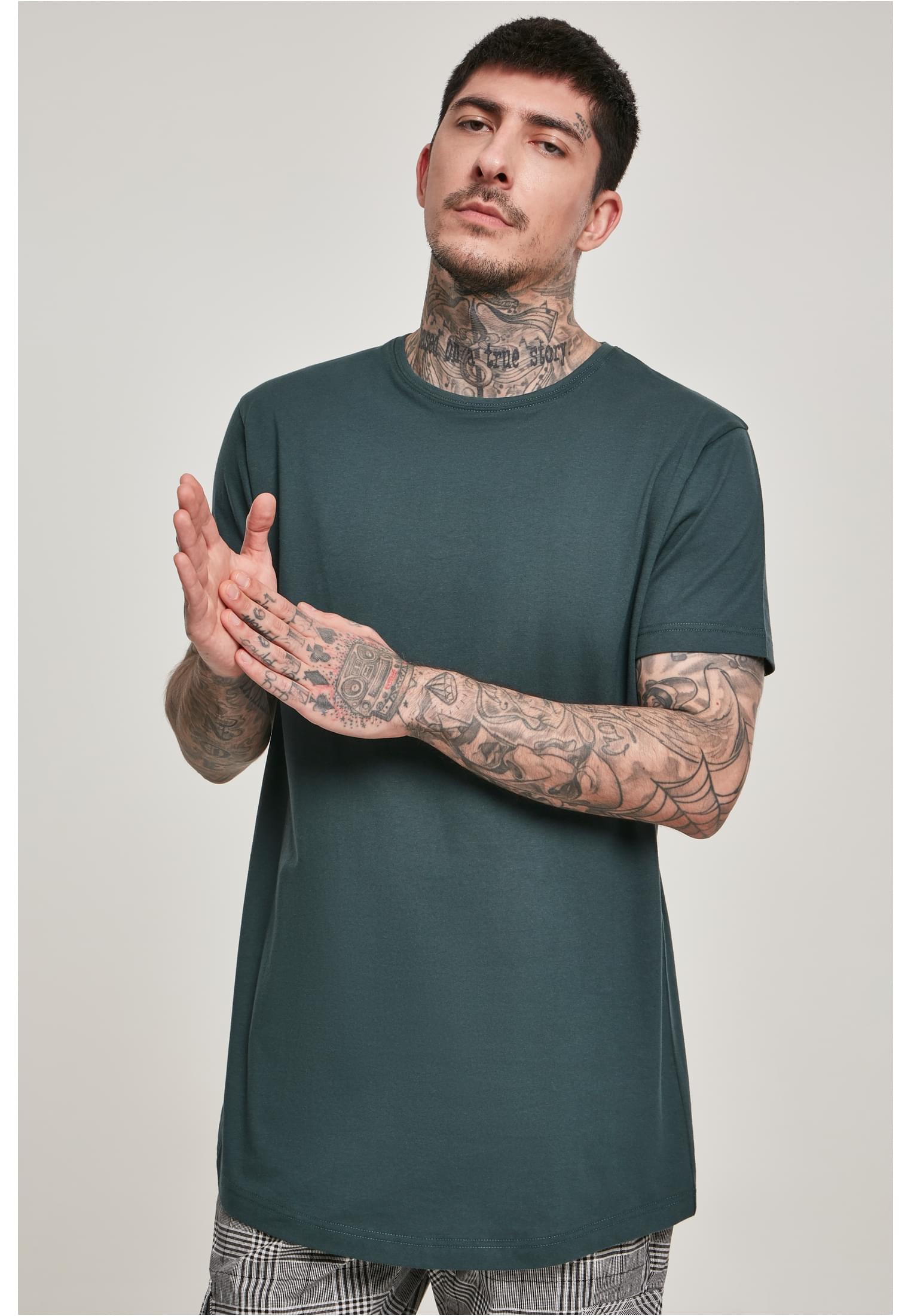 En man med tatueringar på armarna och halsen poserar i en mörkgrön UC Shaped Long Tee.