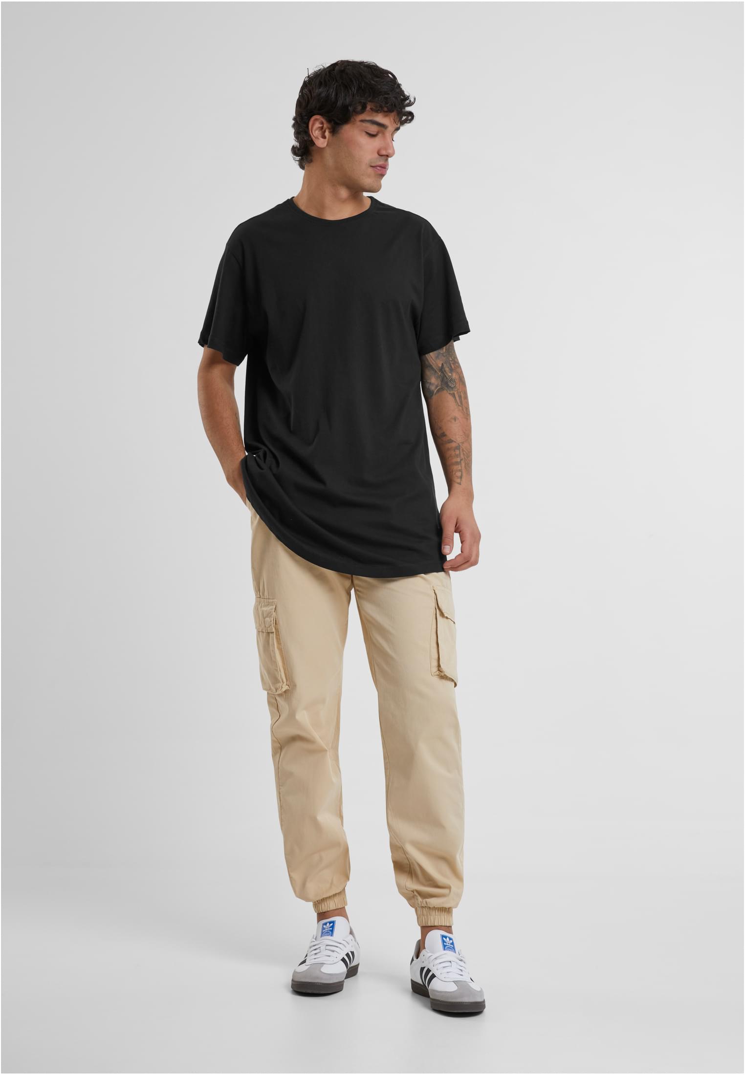En manlig modell som poserar i en svart lång t-shirt med rundad fåll, beiga cargobyxor och vita sneakers mot en ljus bakgrund.