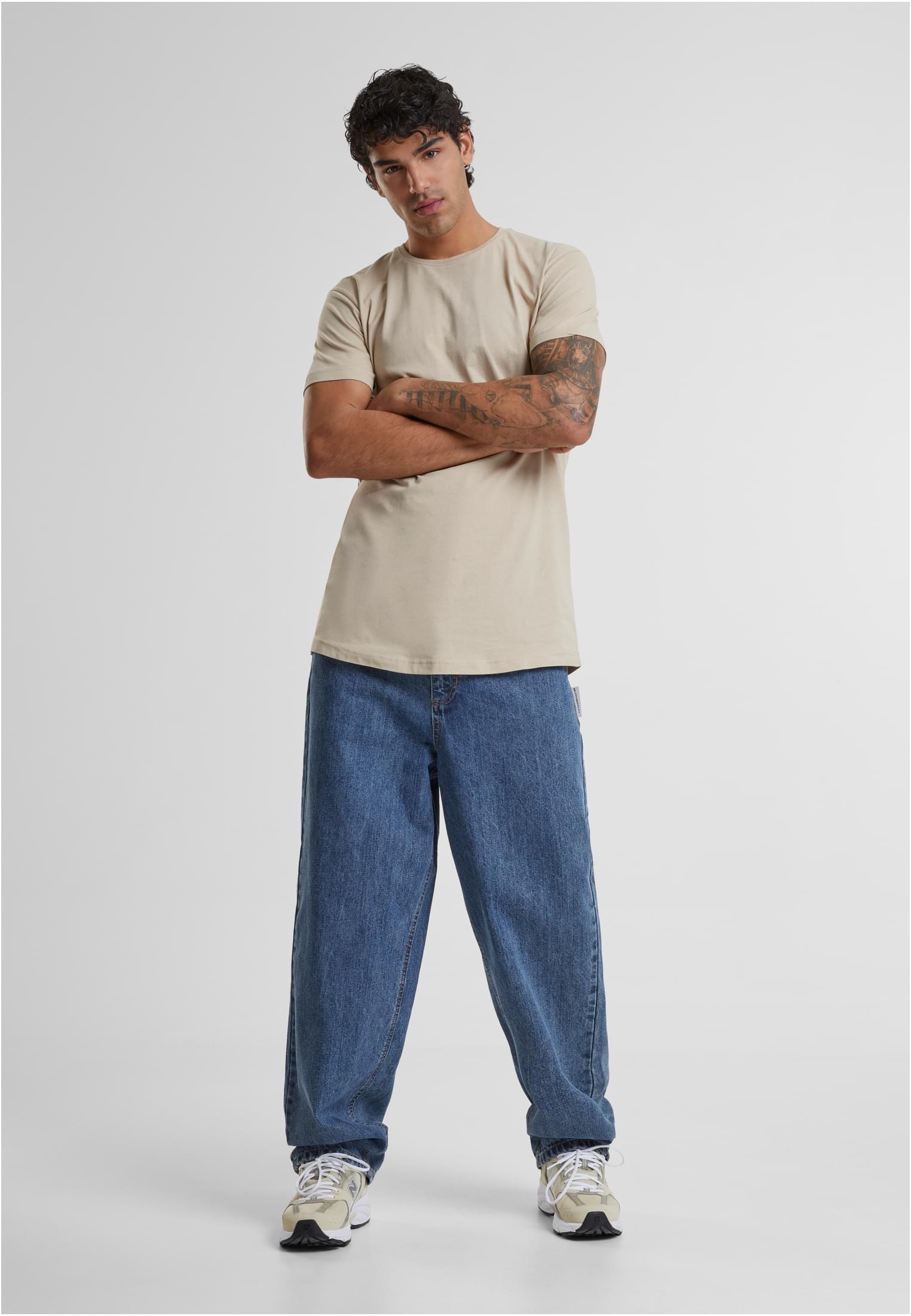 En man som bär en beige Shaped Long Tee med lång passform och vida blåa jeans.