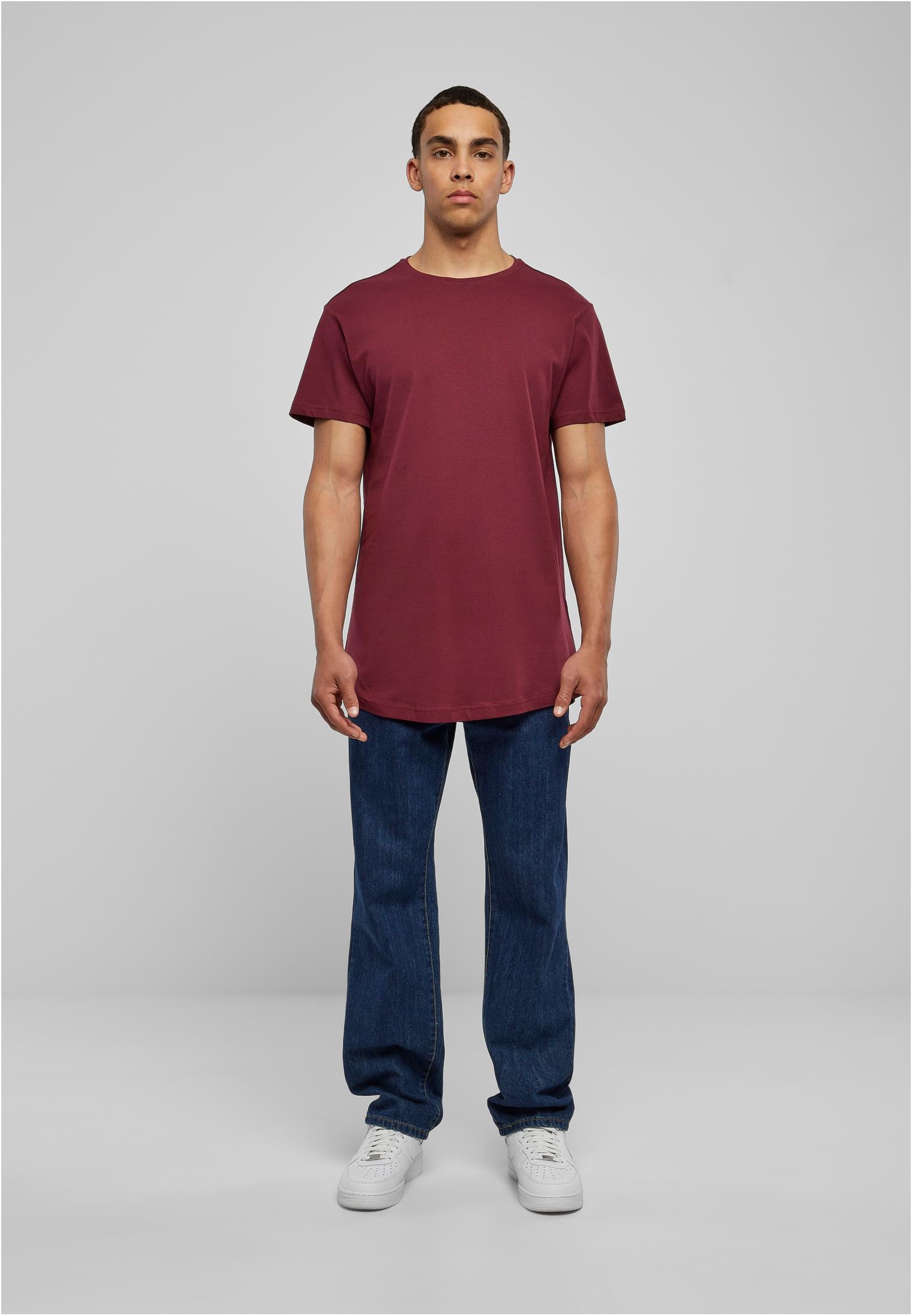 En man poserar i en terrakottafärgad UC Shaped Long Tee, mörkblå jeans och vita sneakers mot en grå bakgrund.