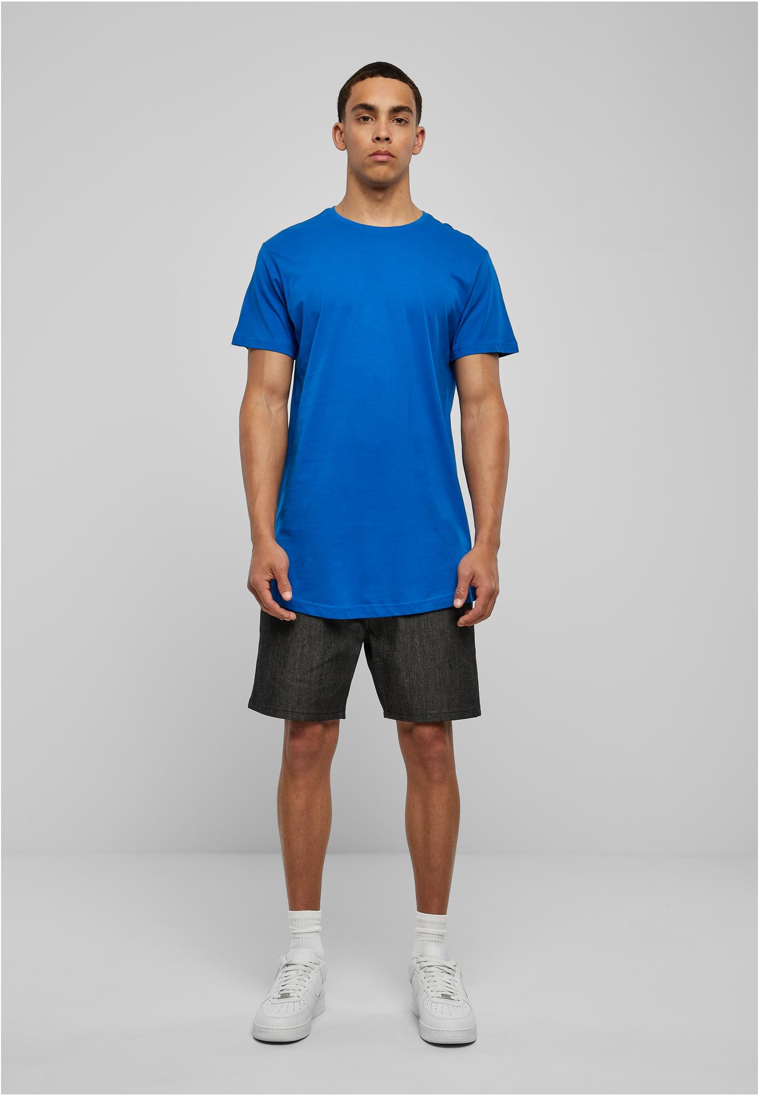 Manlig modell som poserar framifrån i en blå Shaped Long Tee, mörkgrå shorts och vita sneakers mot en ljusgrå bakgrund.