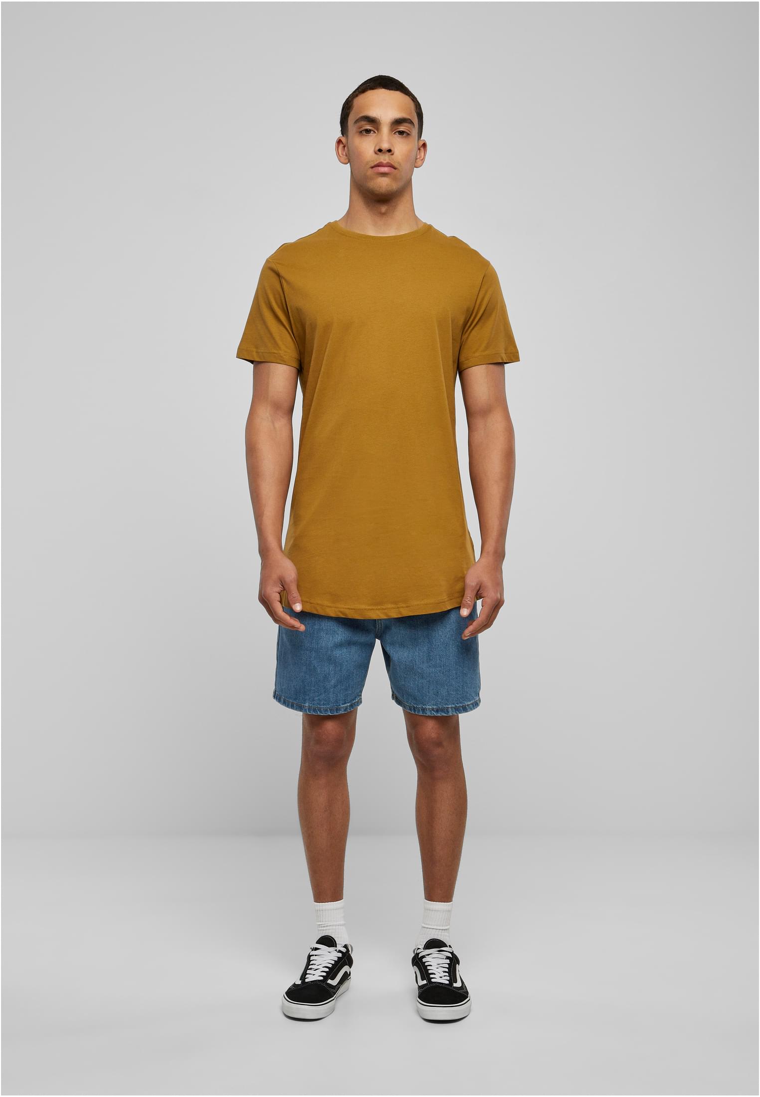 En manlig modell poserar i en terrakottafärgad Shaped Long Tee, blå jeansshorts och svarta sneakers, fotograferad framifrån mot en ljusgrå bakgrund.