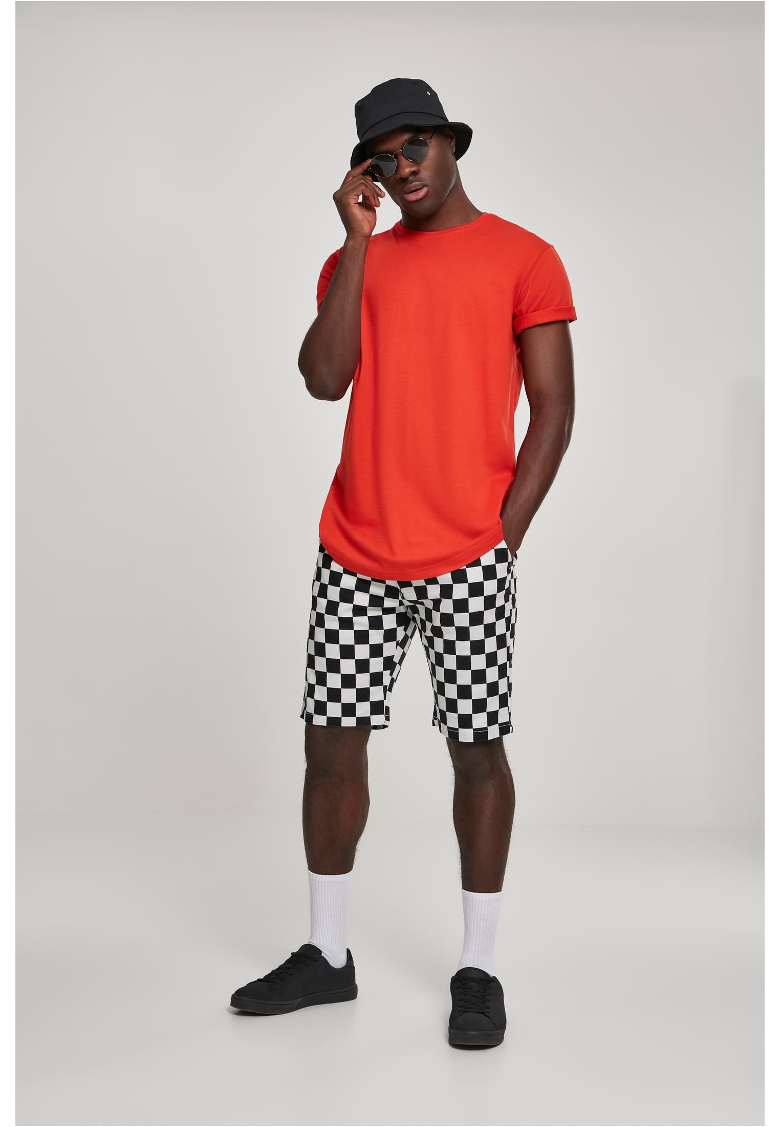 En manlig modell poserar i en terrakottafärgad UC Shaped Long Tee, stylad med schackrutiga shorts, en svart bucket-hatt och solglasögon.