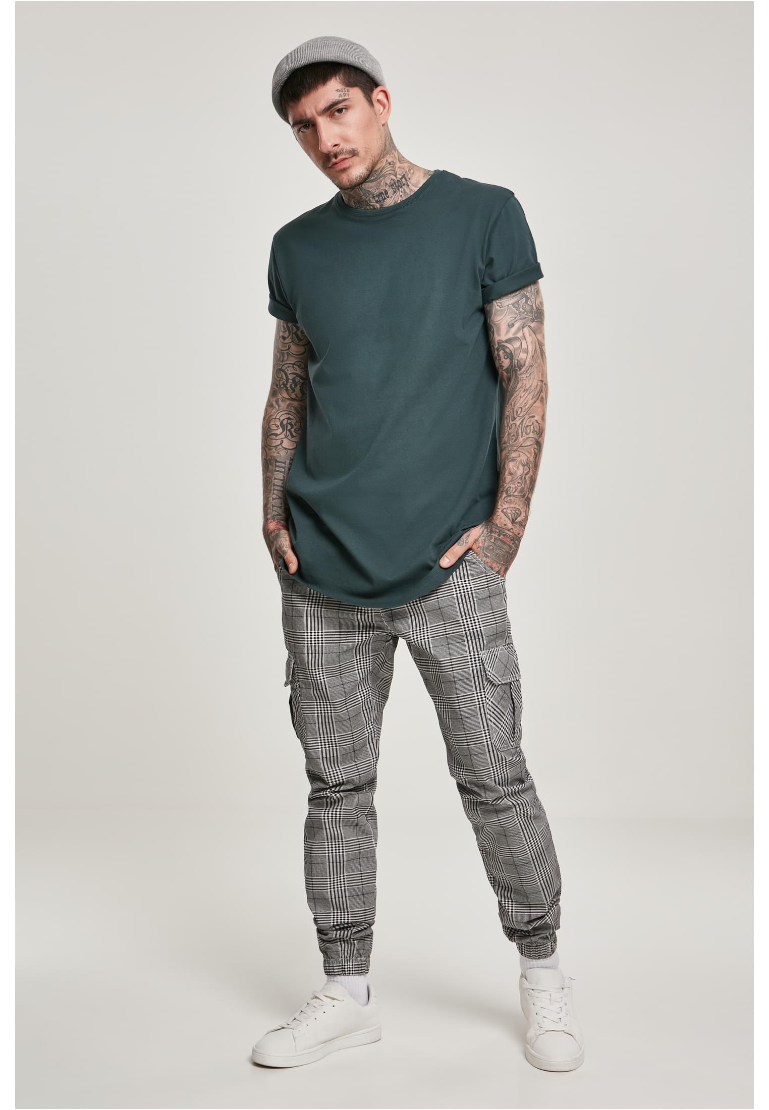 En man med tatueringar som bär en mörkgrön UC Shaped Long Tee, rutiga byxor, vita sneakers och en grå mössa.