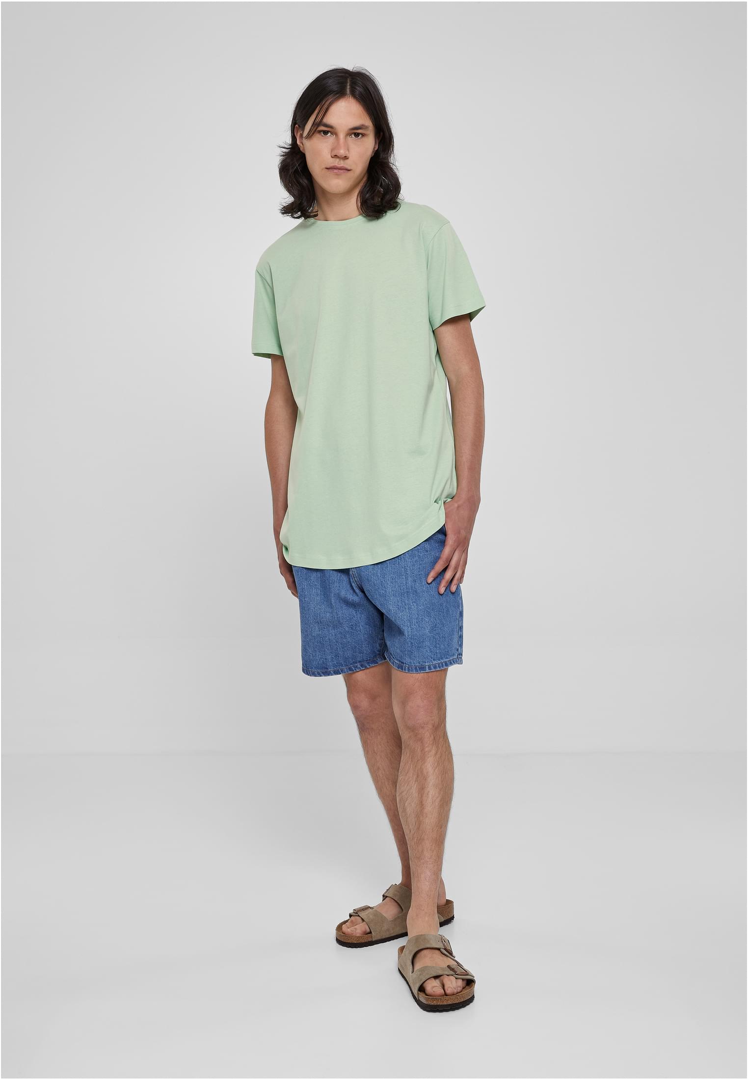 Manlig modell i helfigur som bär en ljusgrön Shaped Long Tee, blåa jeansshorts och sandaler, mot en vit bakgrund.