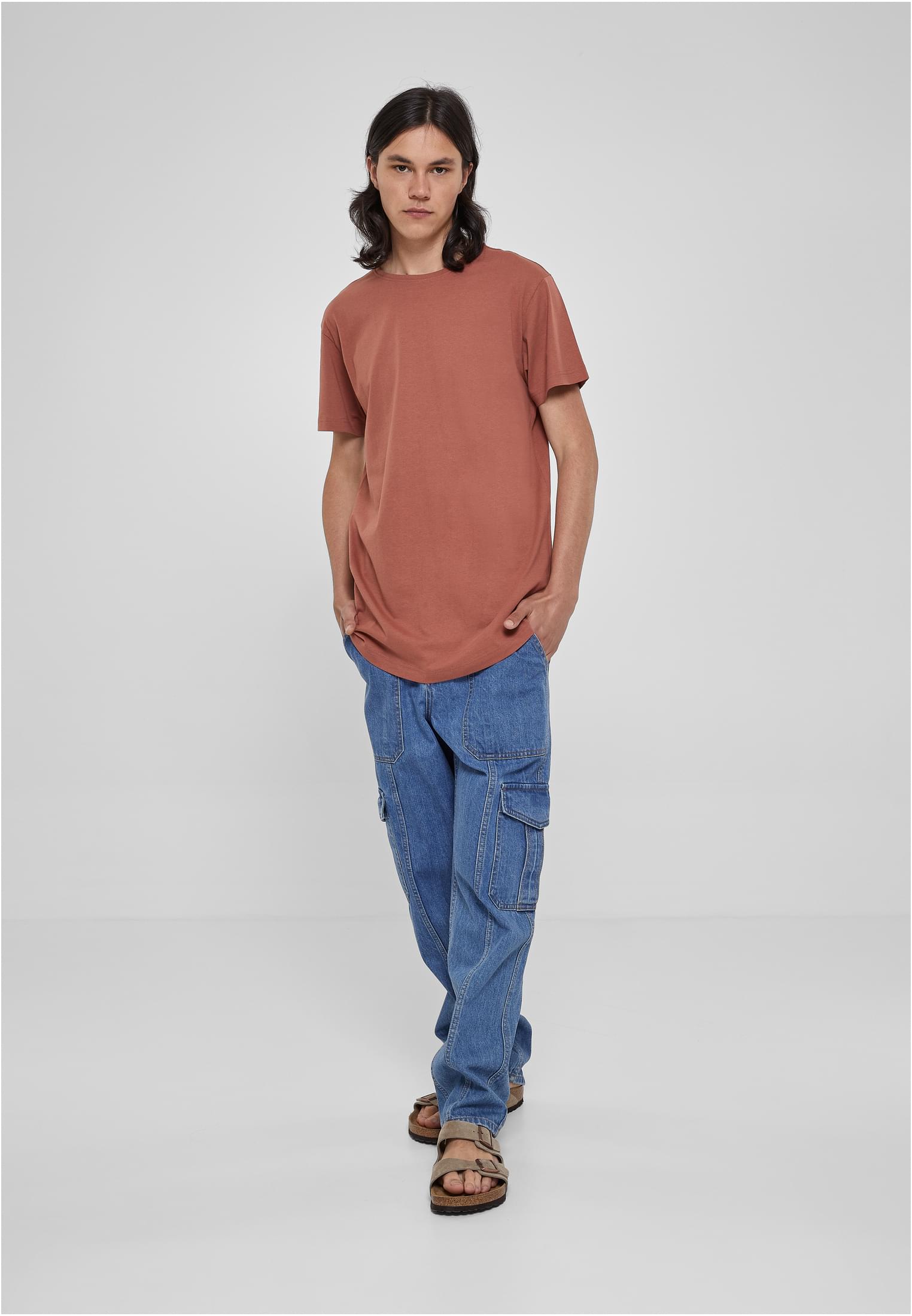 En manlig modell poserar i en terrakottafärgad UC Shaped Long Tee, blå cargobyxor och sandaler. Han står framifrån mot en ljusgrå studiobakgrund.