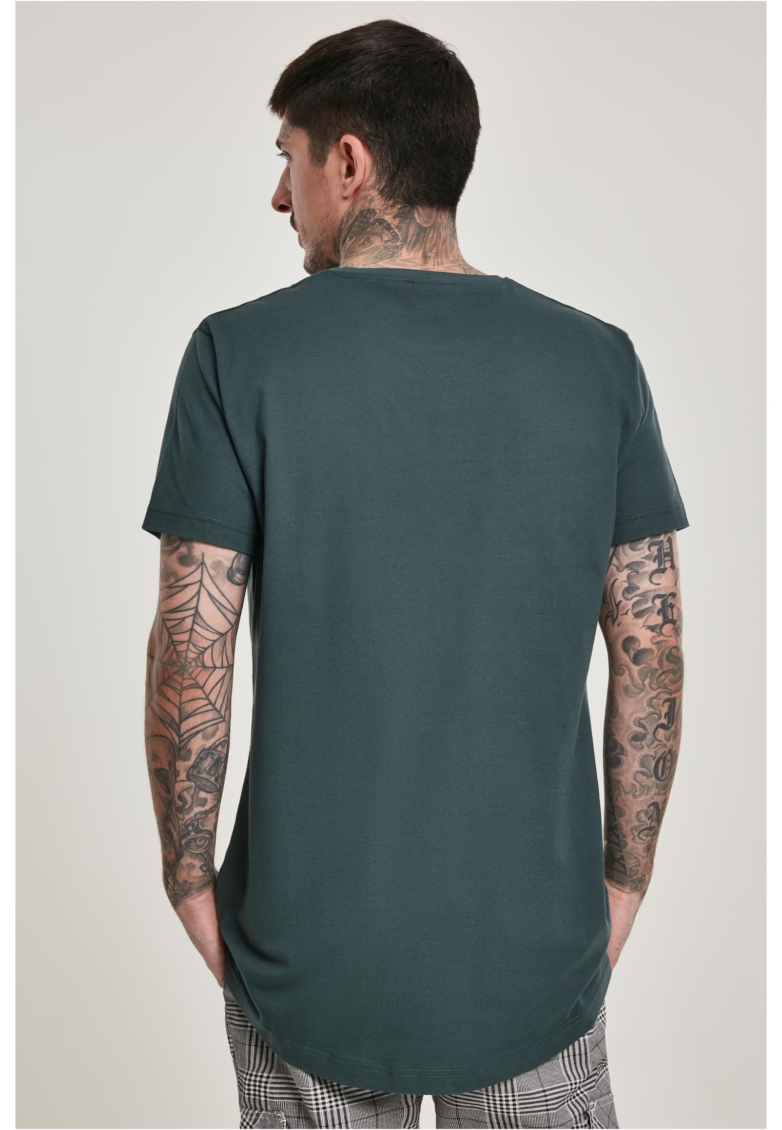 Man med tatueringar sedd bakifrån, klädd i en mörkgrön UC Shaped Long Tee med en längre passform och rundad nederkant.