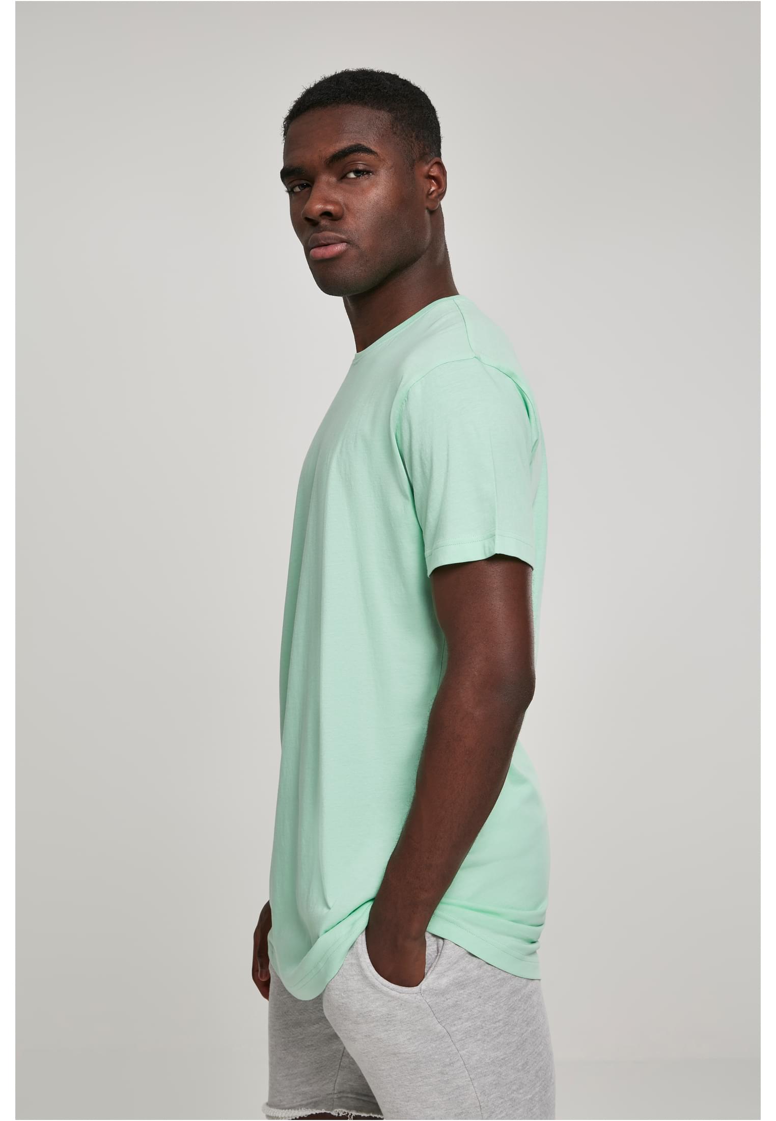 Man som bär en ljusgrön UC Shaped Long Tee och grå shorts, fotograferad snett från sidan mot en ljusgrå bakgrund.