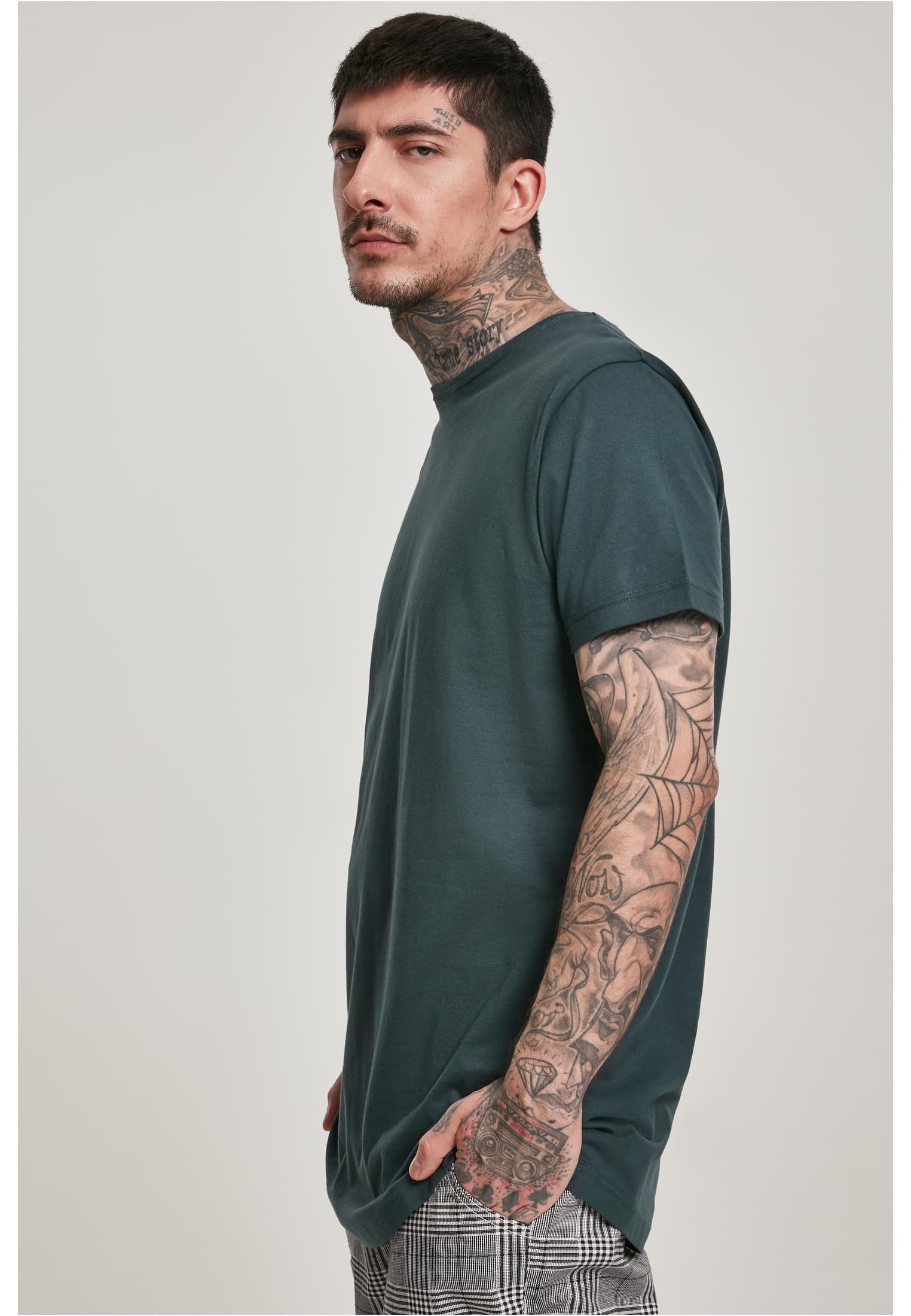 En man med tatueringar på armar och hals bär en mörkgrön UC Shaped Long Tee, fotograferad snett framifrån mot en ljusgrå bakgrund.