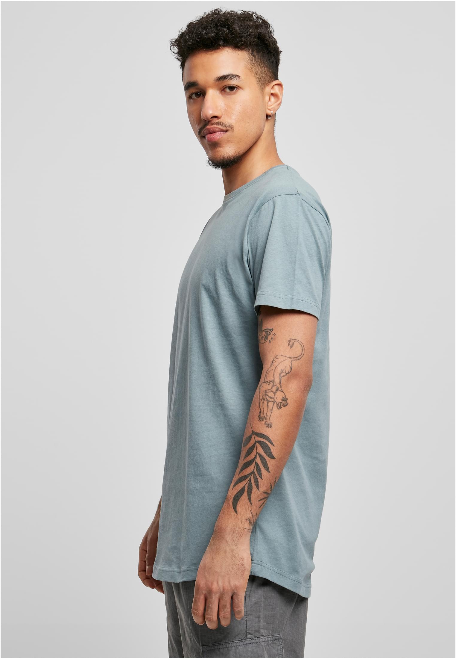 Manlig modell i en blågrön UC Shaped Long Tee, fotograferad i trekvartsprofil mot en ljusgrå bakgrund.