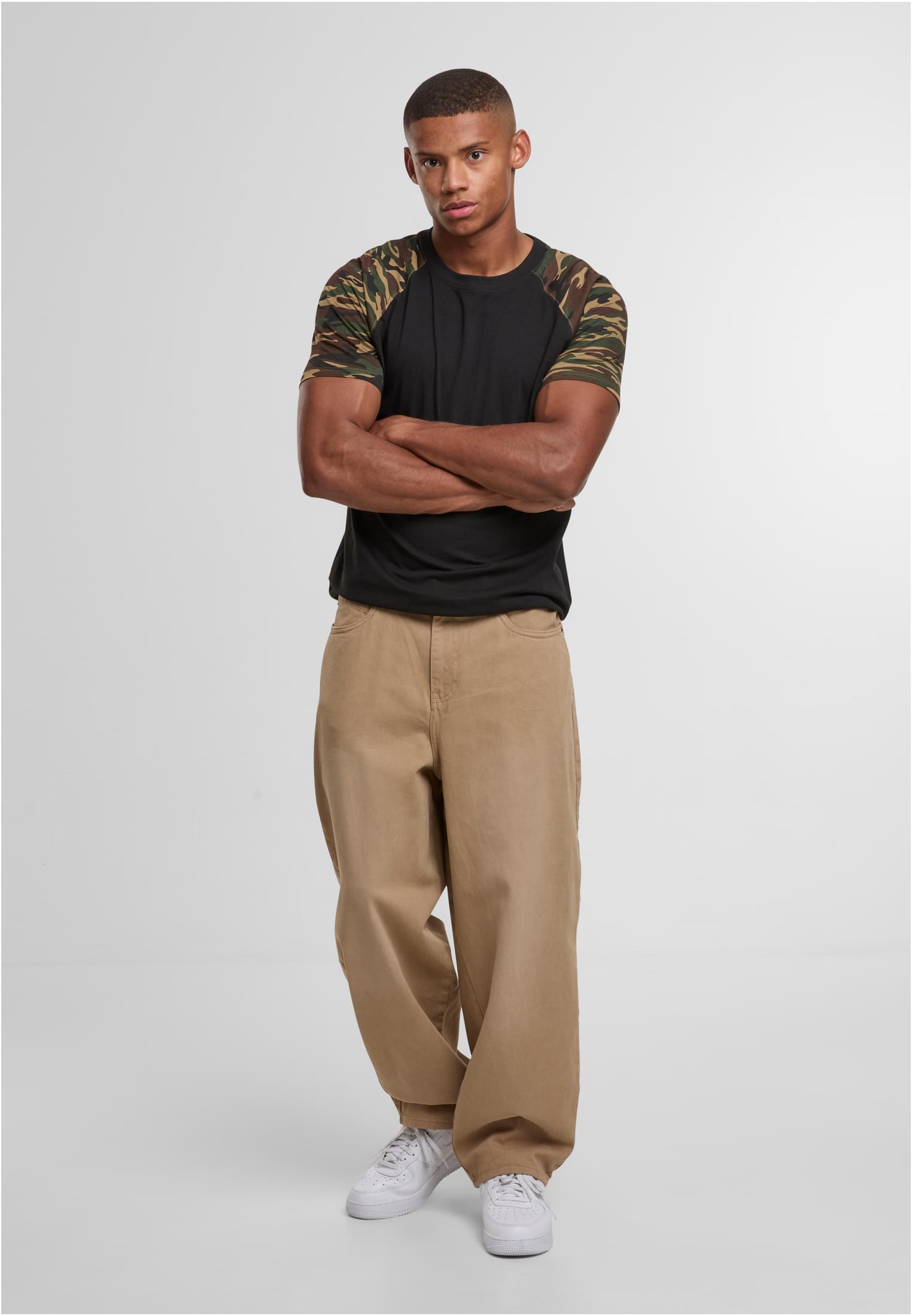 En manlig modell poserar med armarna i kors i en svart raglan-t-shirt med kamouflagemönstrade ärmar, stylad med vida beigefärgade byxor och vita sneakers.