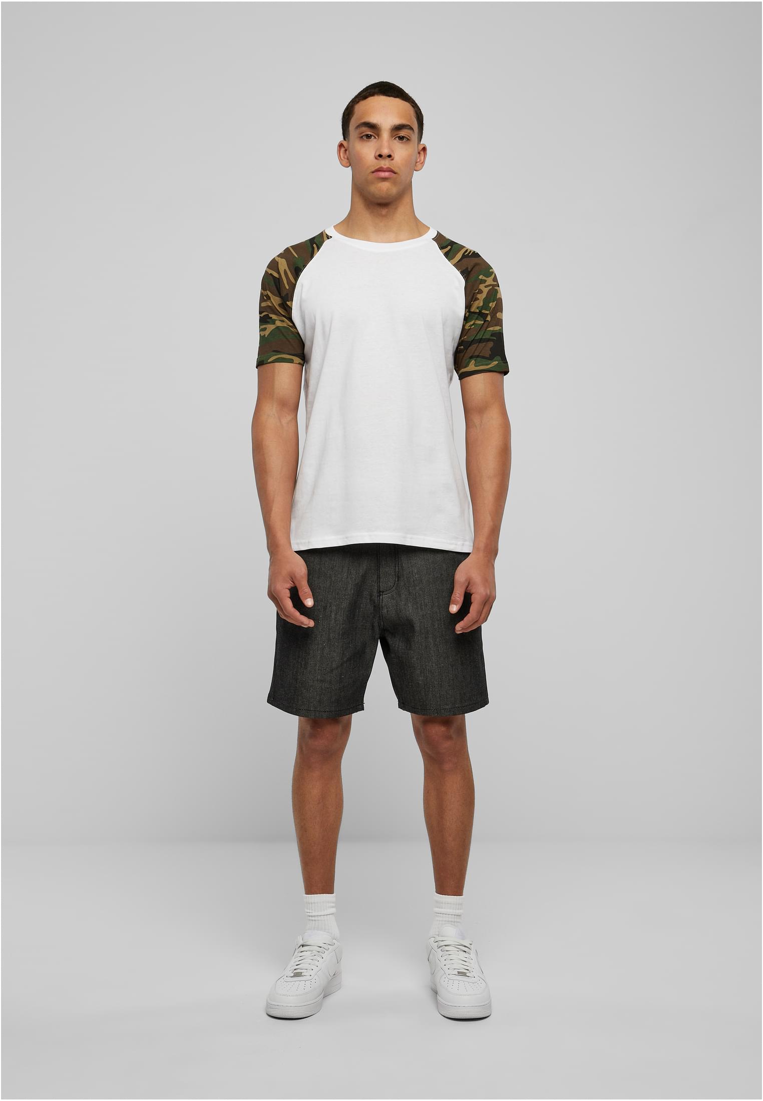 En man fotograferad framifrån i en vit Raglan Contrast Tee med kamouflagemönstrade ärmar, mörka shorts och vita sneakers.