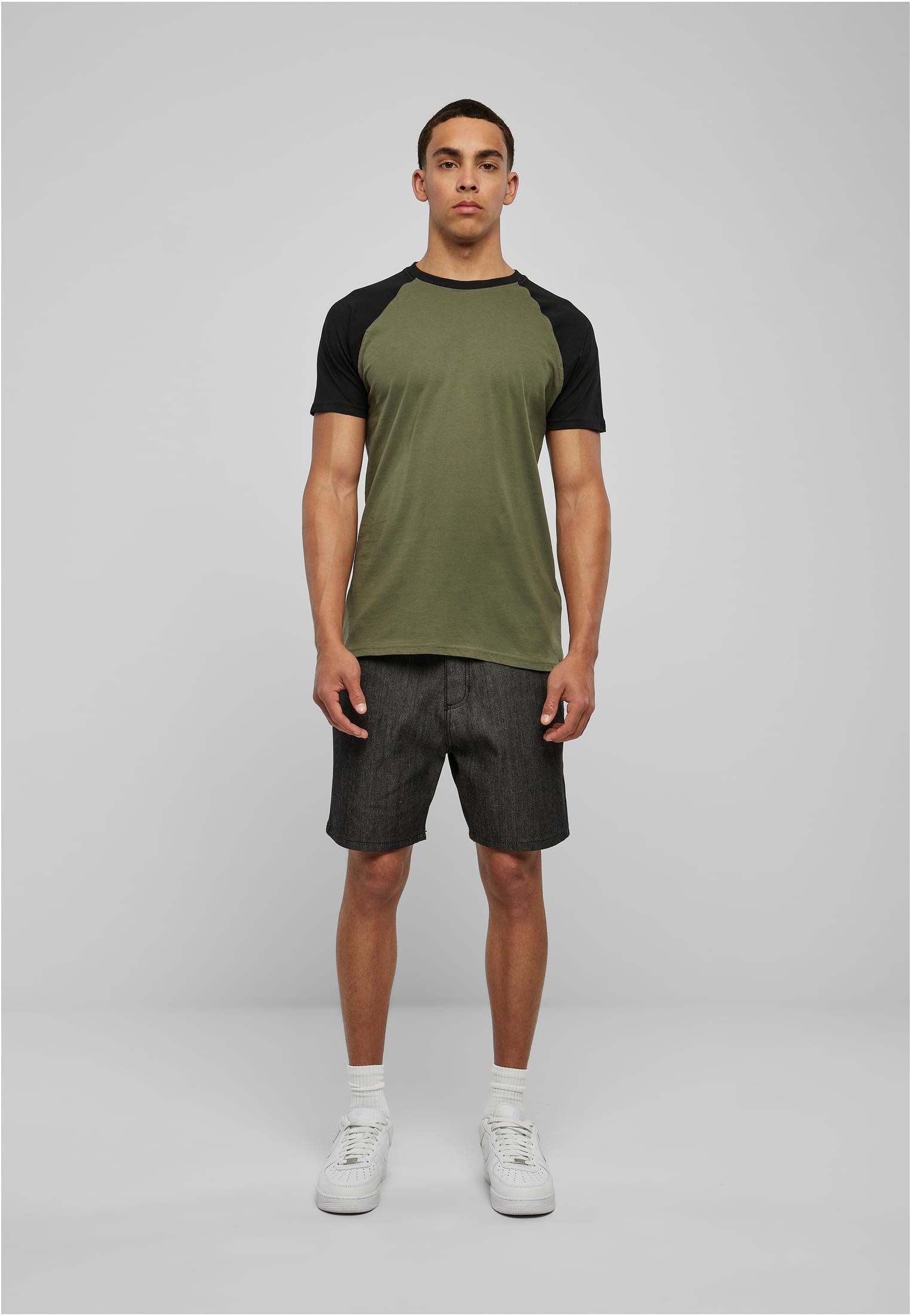 En manlig modell sedd framifrån som bär en tvåfärgad raglan-t-shirt med grön kropp och svarta ärmar, mörka shorts och vita sneakers.