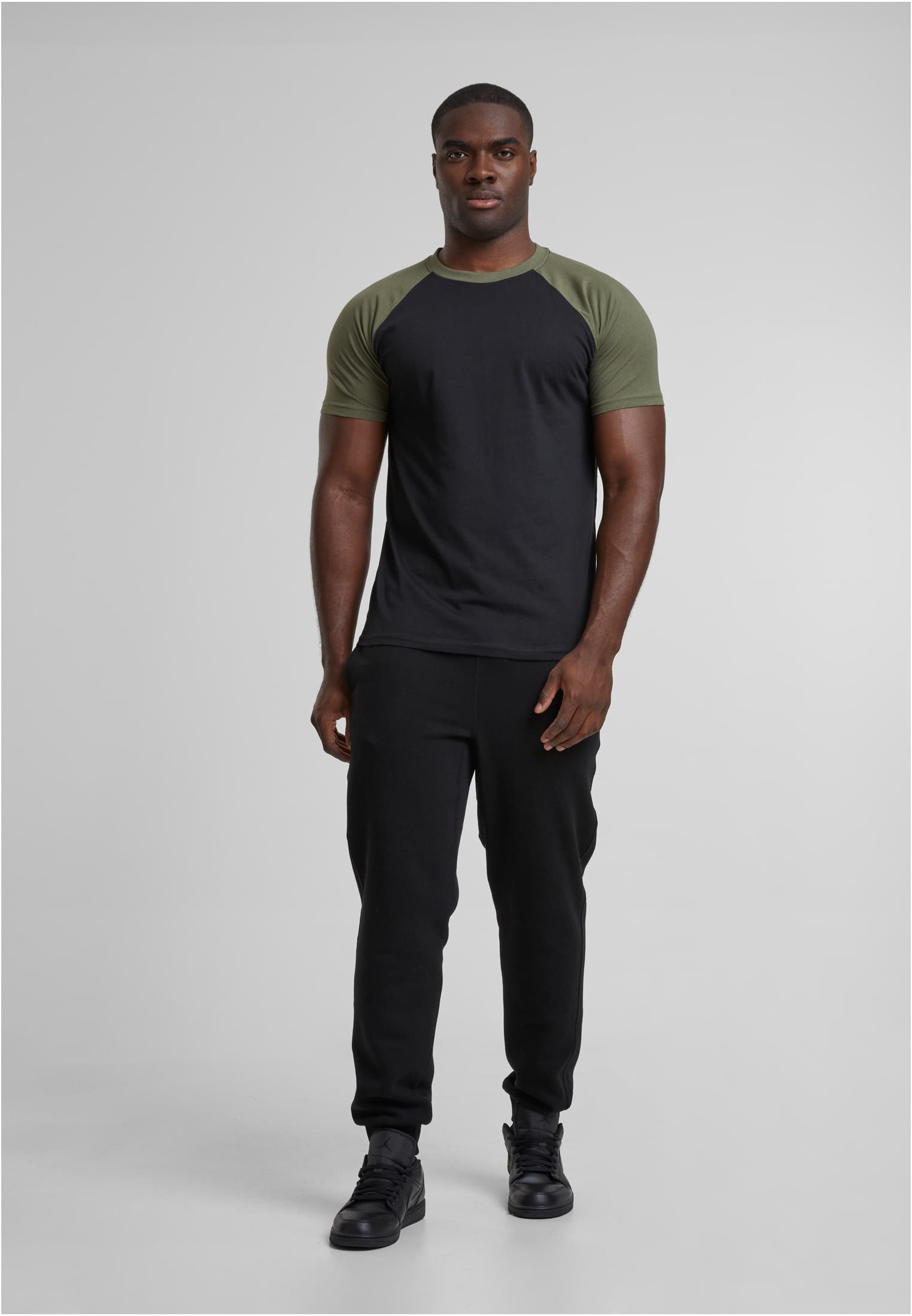 En man poserar i en svart t-shirt med olivgröna raglanärmar och svarta byxor.