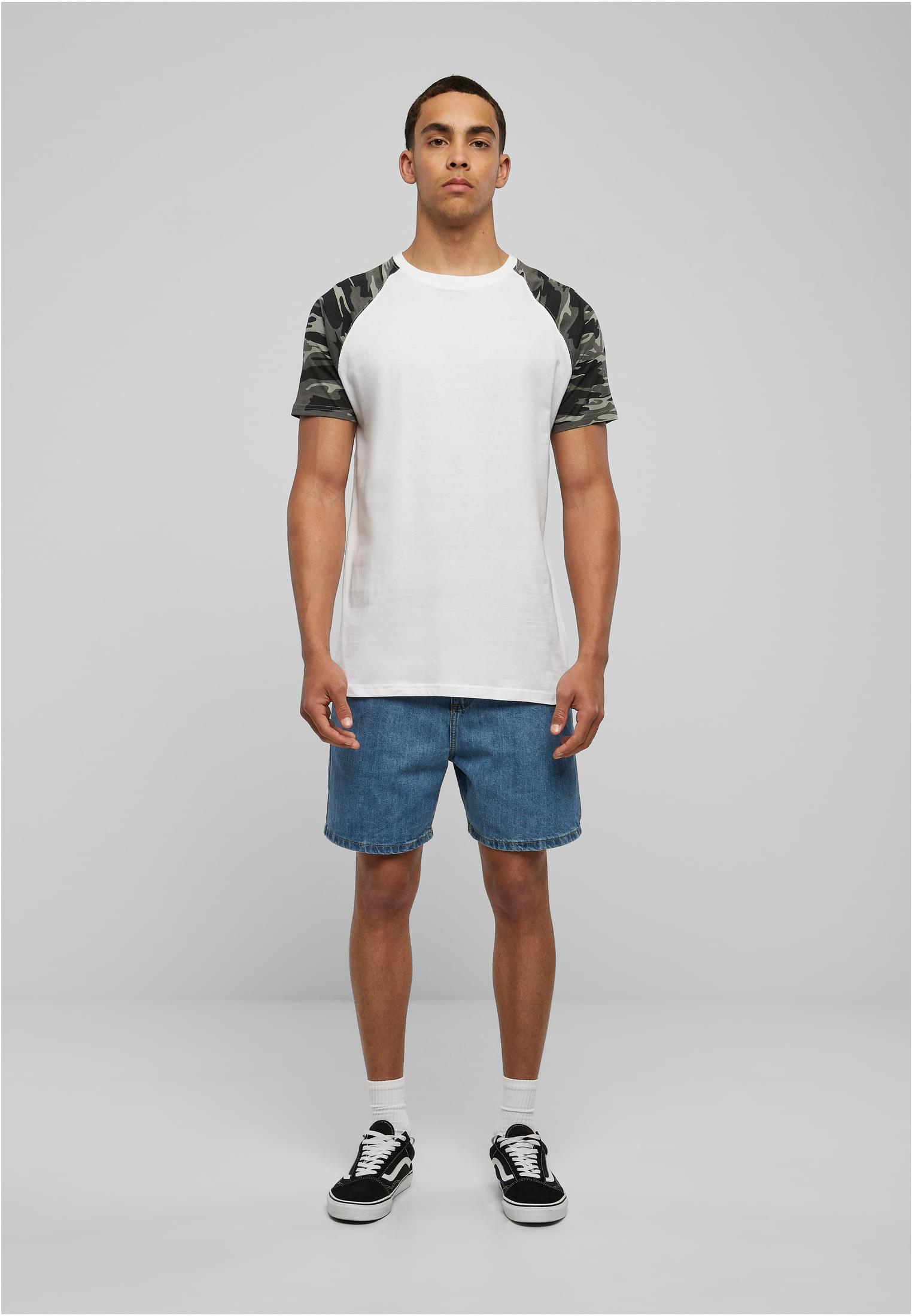 Helkroppsbild framifrån av en manlig modell som bär en vit raglan-t-shirt med kamouflagemönstrade ärmar, blå jeansshorts och svartvita sneakers.