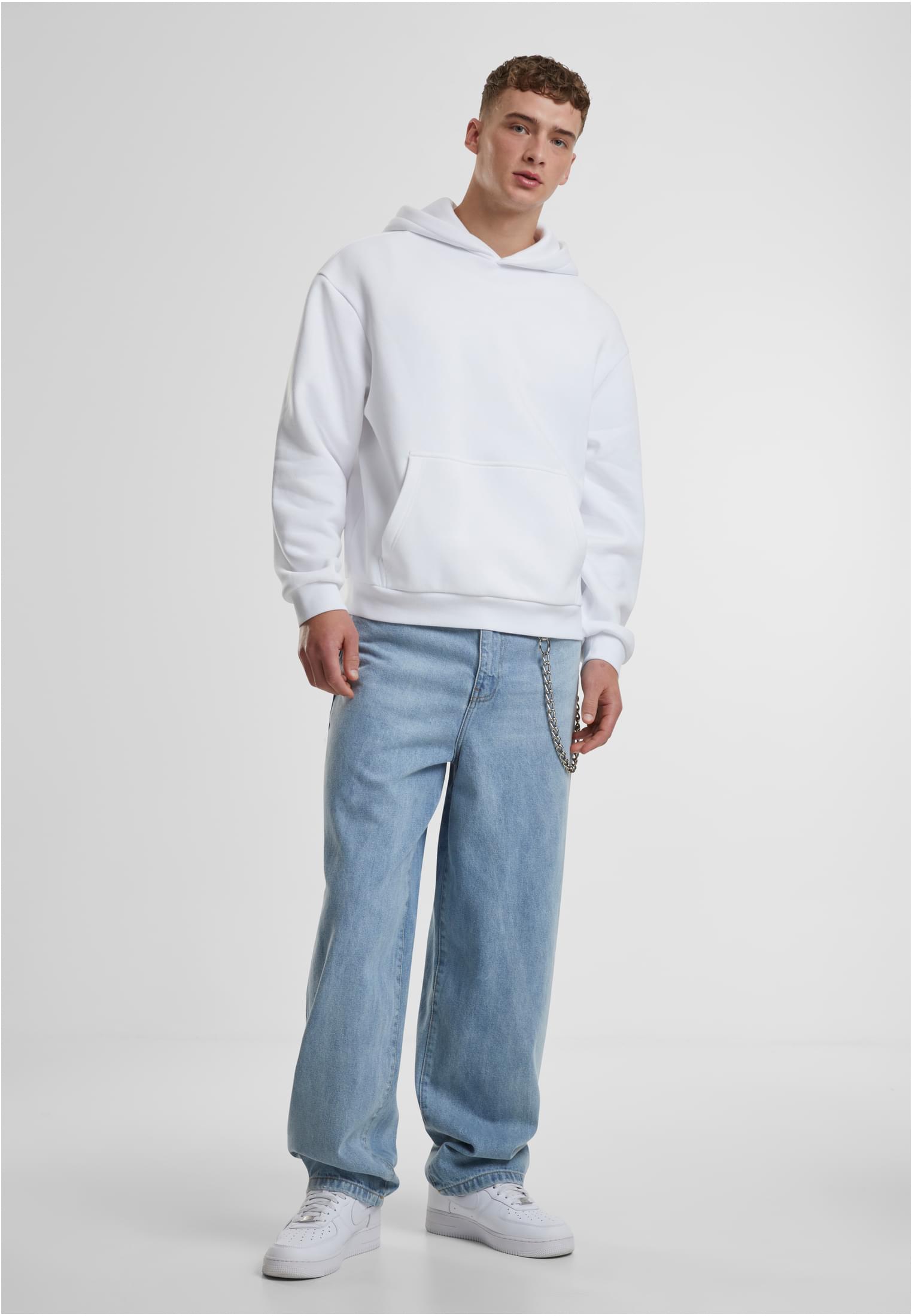 En man poserar i en vit luvtröja, ljusblå baggy jeans med en kedja och vita sneakers. Han är fotograferad rakt framifrån i en studio.