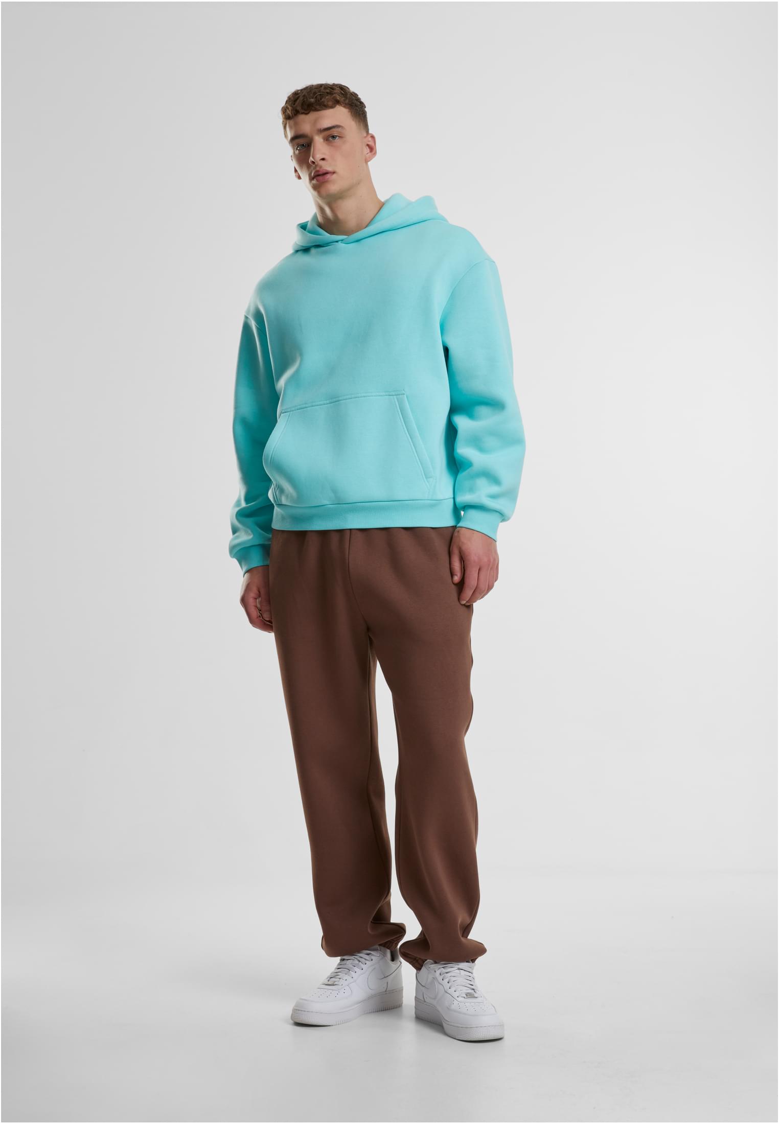 En man i en ljusblå Fluffy Hoody och bruna byxor står framför en vit bakgrund.
