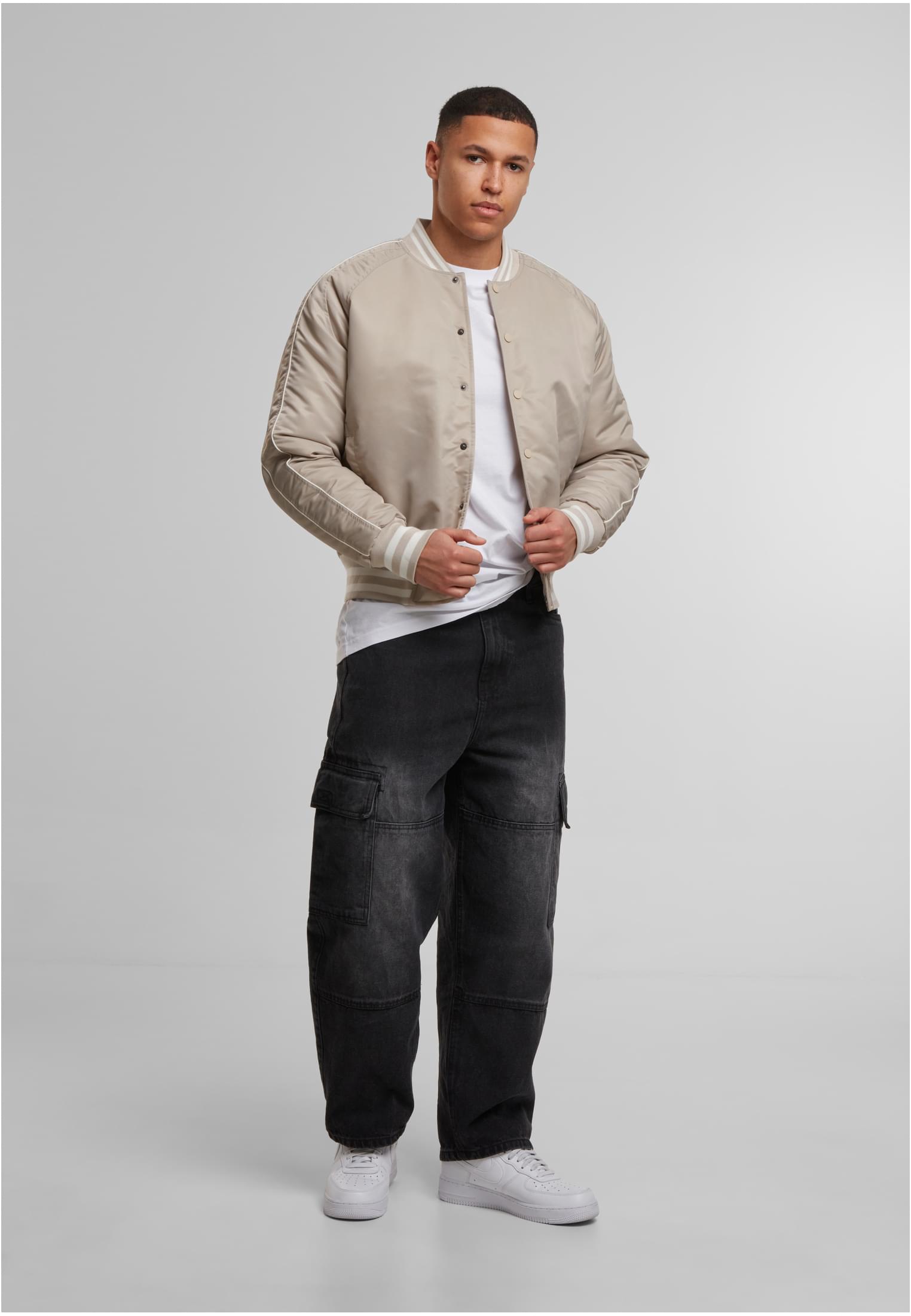 En man visar framsidan av en beige UC Racer Bomber Jacket, stylad med en vit t-shirt och svarta cargobyxor.