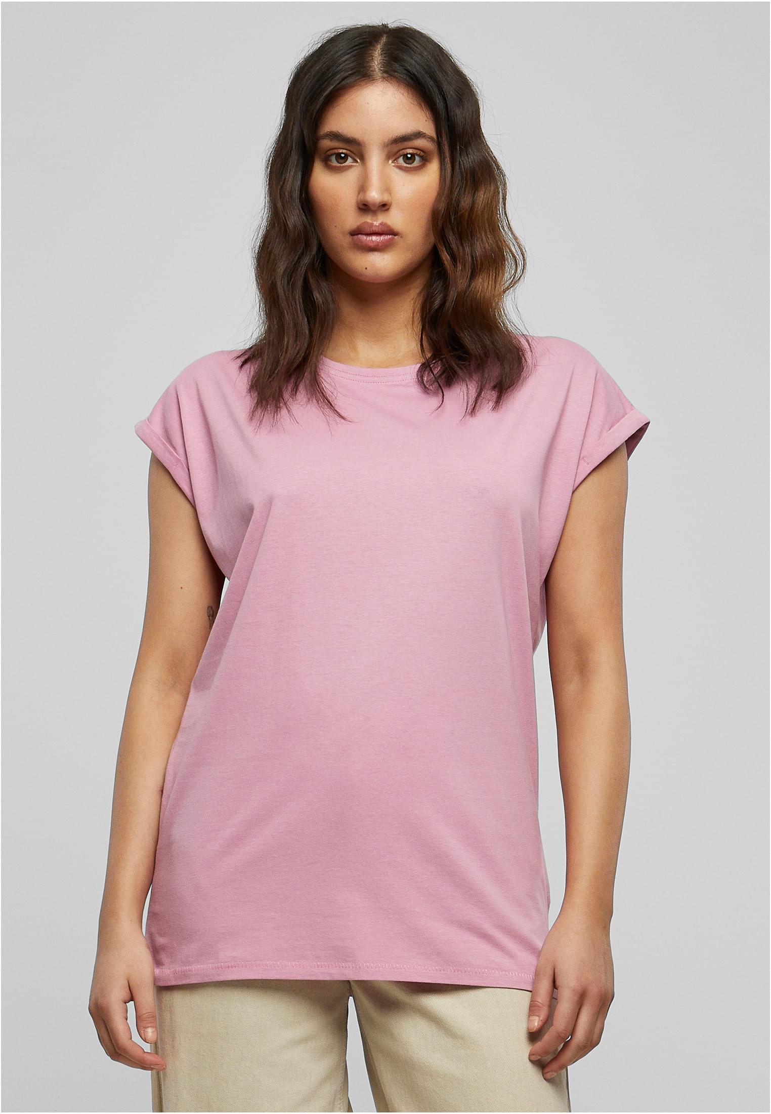 En kvinnlig modell som bär en ljusrosa UC Ladies Extended Shoulder Tee. Hon är fotograferad framifrån mot en ljusgrå bakgrund.