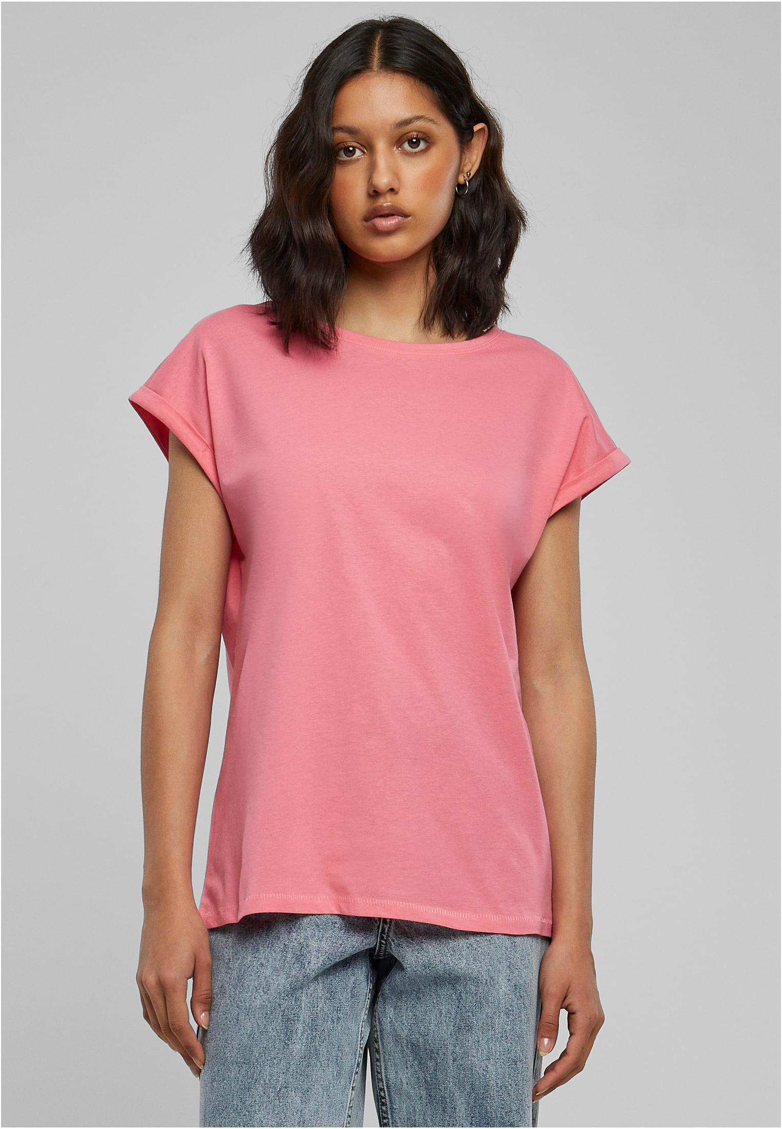 En kvinna sedd framifrån, klädd i en rosa t-shirt med nedhasade axlar och uppvikta ärmar, tillsammans med jeans.
