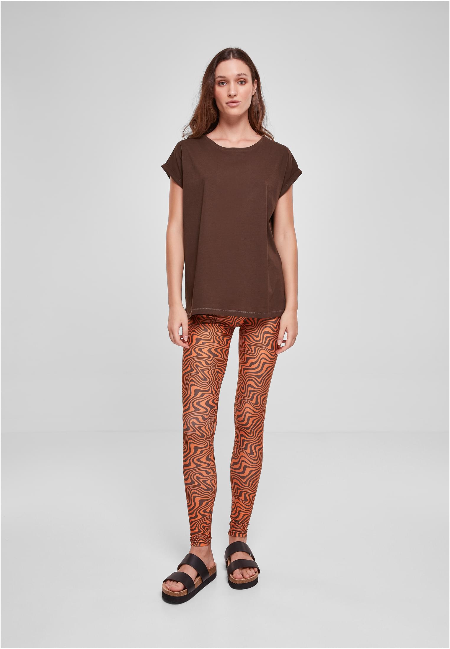 En modell som bär en brun t-shirt med förlängda axlar, orangea och bruna mönstrade leggings och svarta sandaler, fotograferad framifrån.