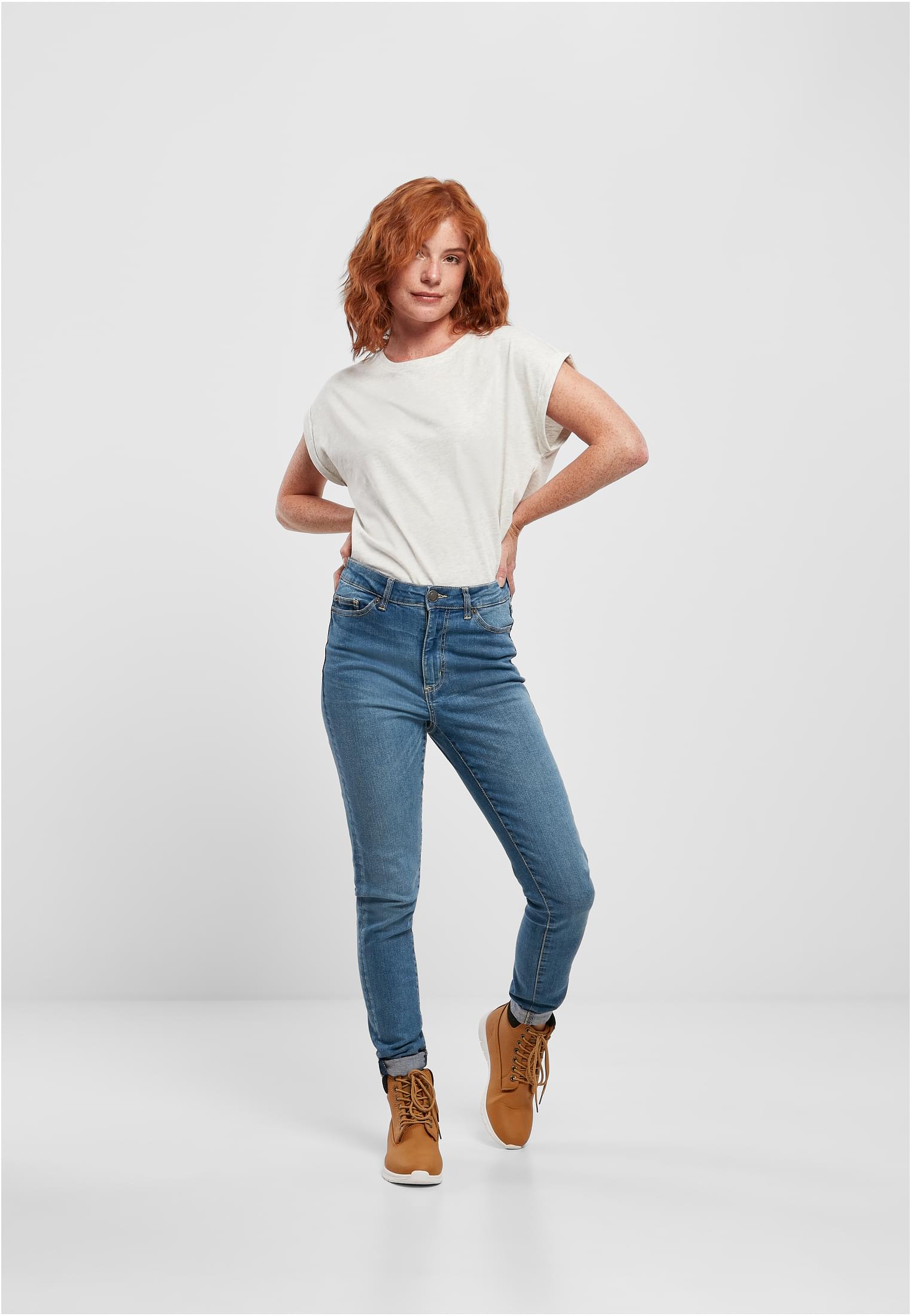 Helkroppsbild av en kvinna med rött hår som bär en ljus Ladies Extended Shoulder Tee, blå jeans och bruna kängor.