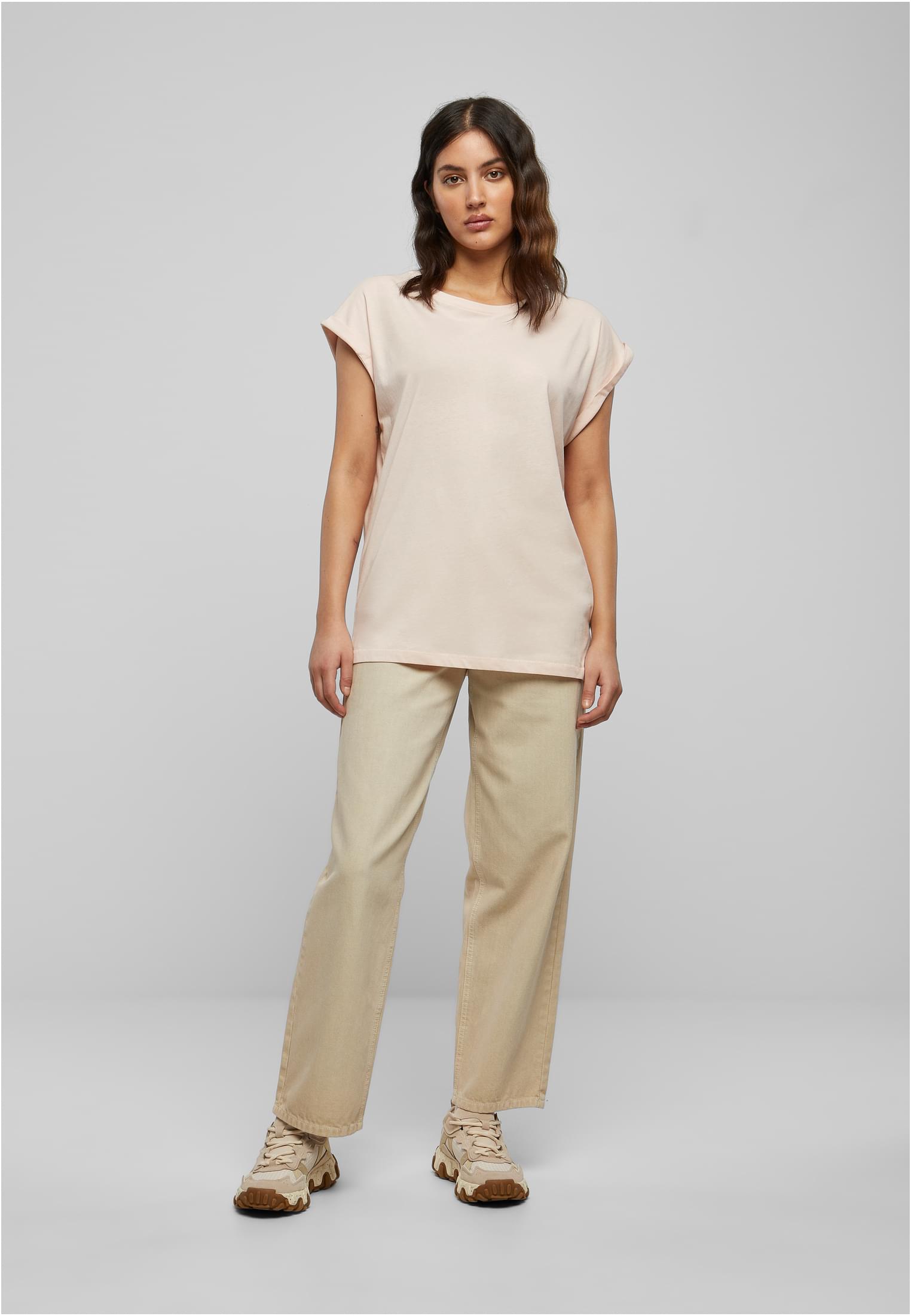 Helbild av en kvinna som bär en ljusrosa t-shirt med förlängd axel, beiga byxor och sneakers.