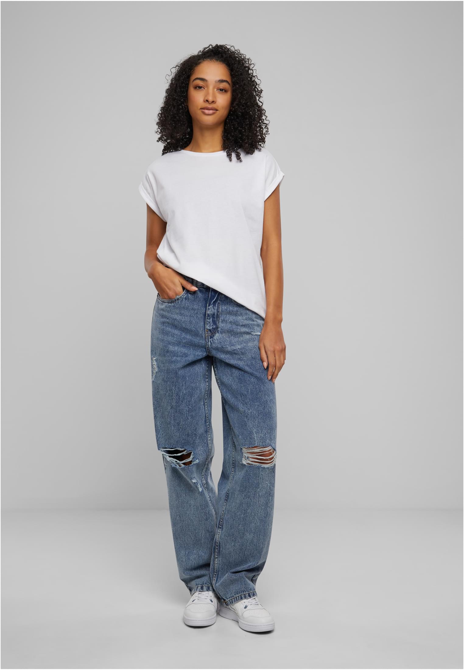 En kvinnlig modell som bär en vit UC Ladies Extended Shoulder Tee med slitna blå jeans och vita sneakers.