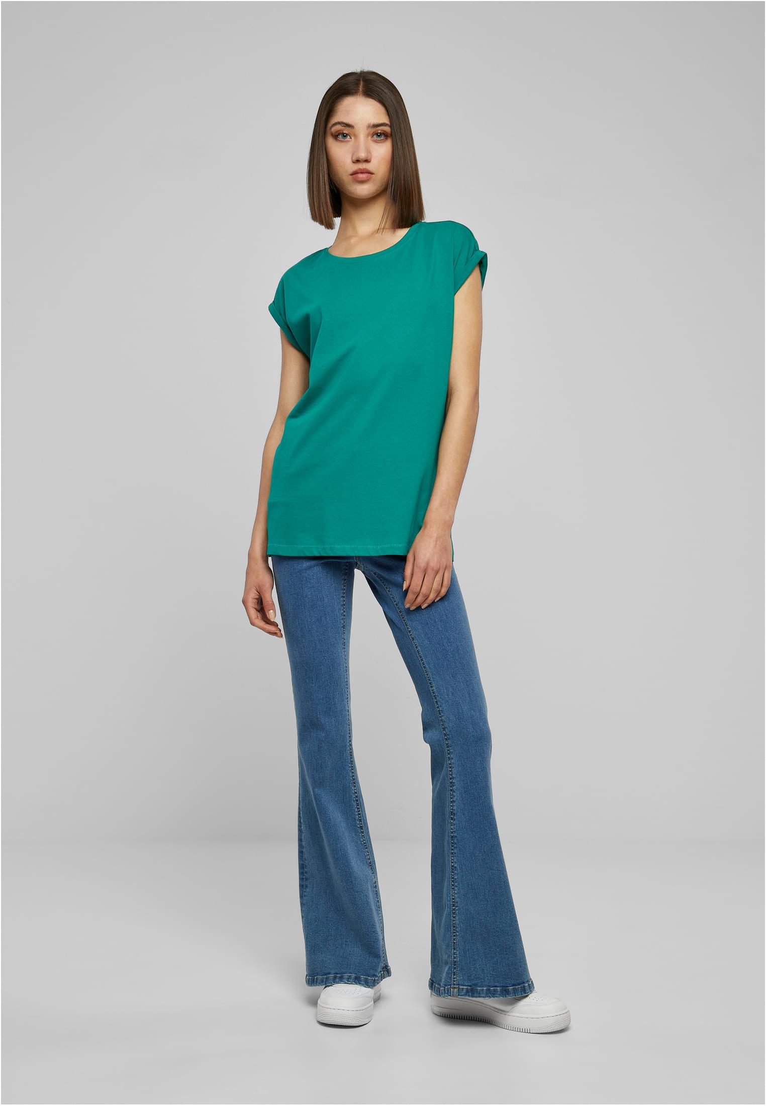 Kvinna i helfigur som bär en grön t-shirt med nedhasade axlar, utsvängda blåjeans och vita sneakers mot en ljusgrå bakgrund.