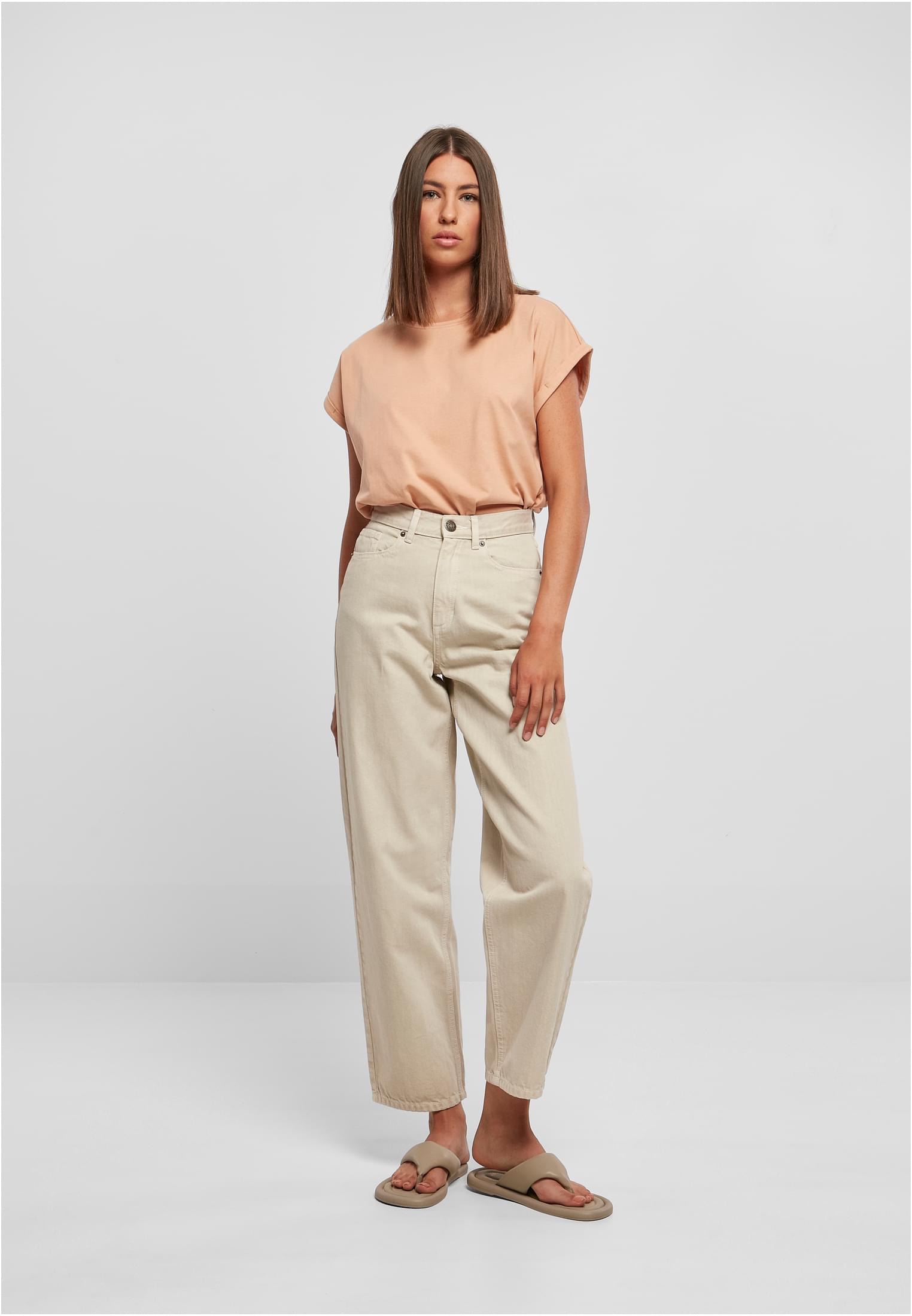 En kvinna fotograferad framifrån i en ljusaprikosfärgad t-shirt med nedhasade axlar, beiga jeans med vida ben och sandaler.