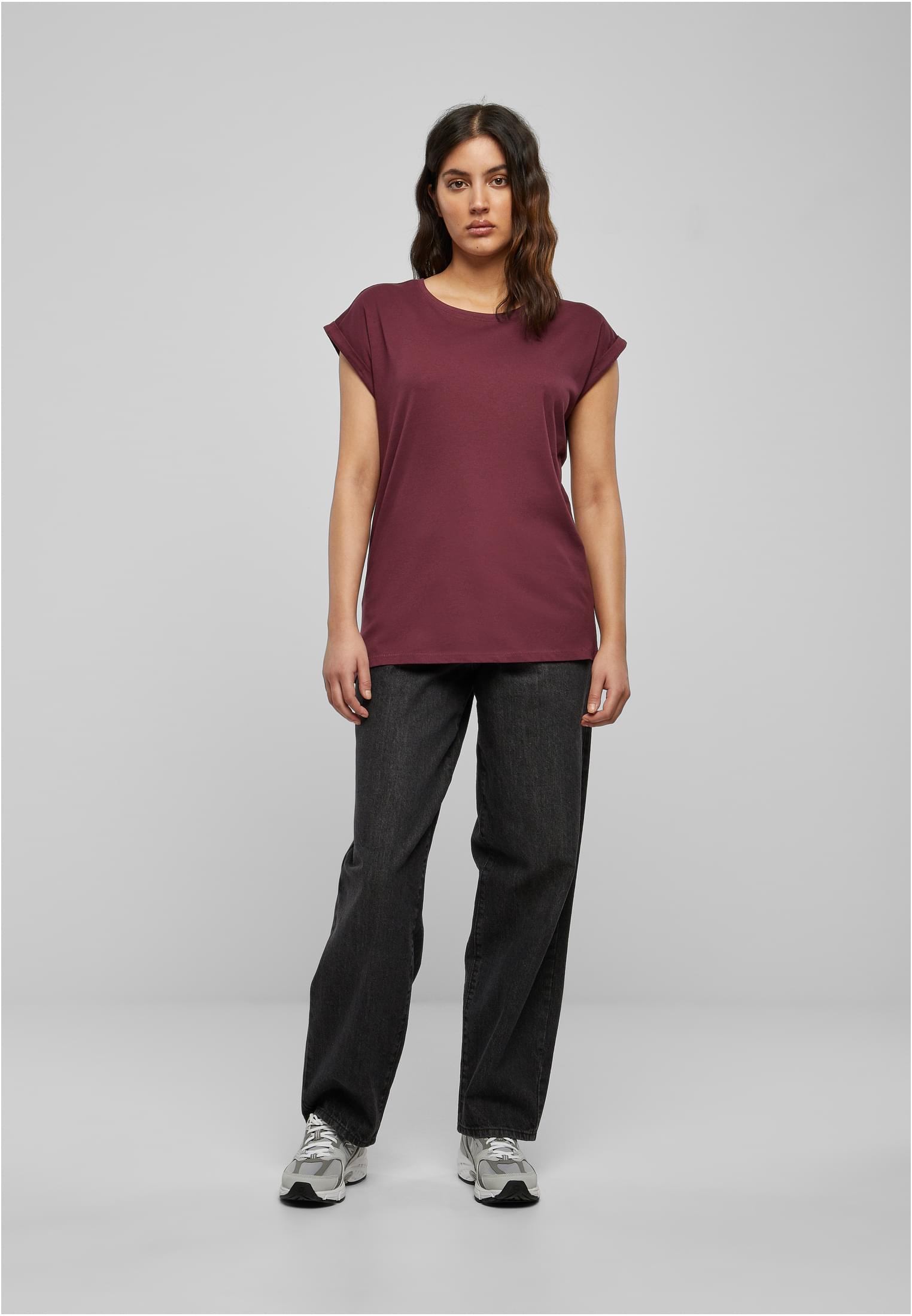En kvinna som bär en vinröd UCL Ladies Extended Shoulder Tee med mörkgråa jeans och sneakers, fotograferad mot en ljusgrå bakgrund.
