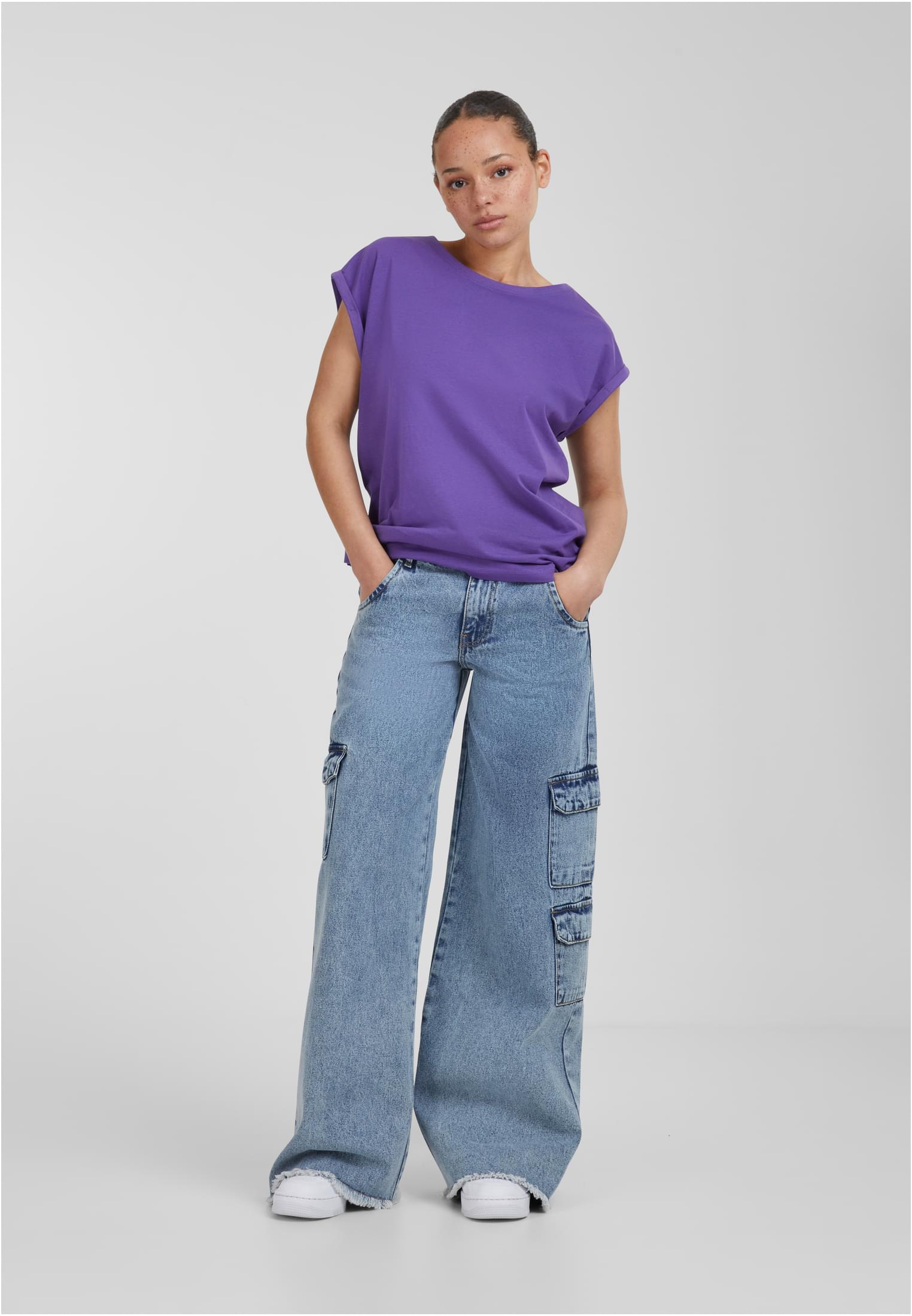 En helkroppsbild av en kvinna som bär en lila t-shirt med nedhasade axlar och vida cargobyxor i ljusblå denim.