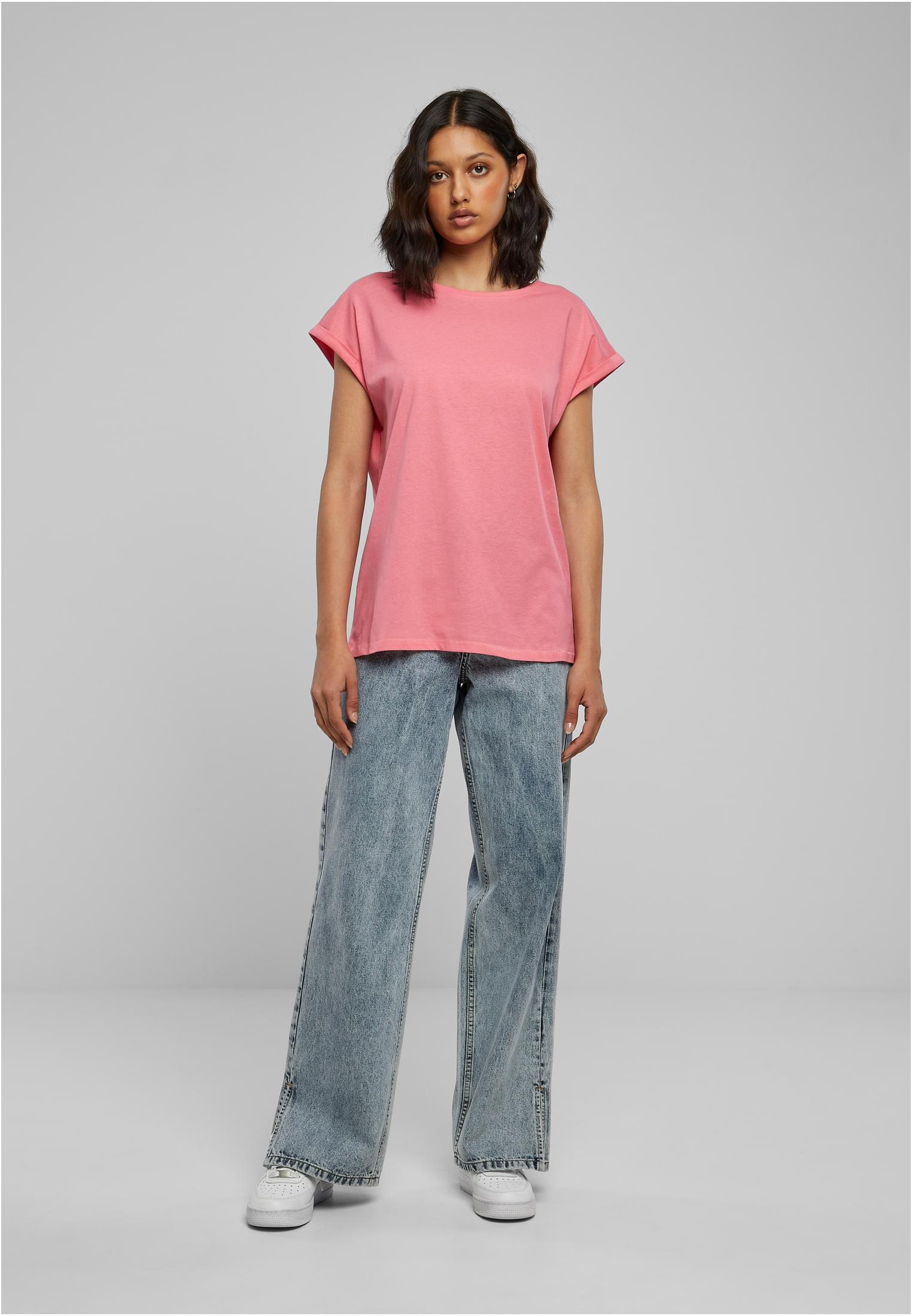 En kvinnlig modell visar upp en rosa t-shirt med nedhasade axlar och uppvikta ärmar, stylad med vida ljusblå jeans och vita sneakers, framför en grå studiobakgrund.