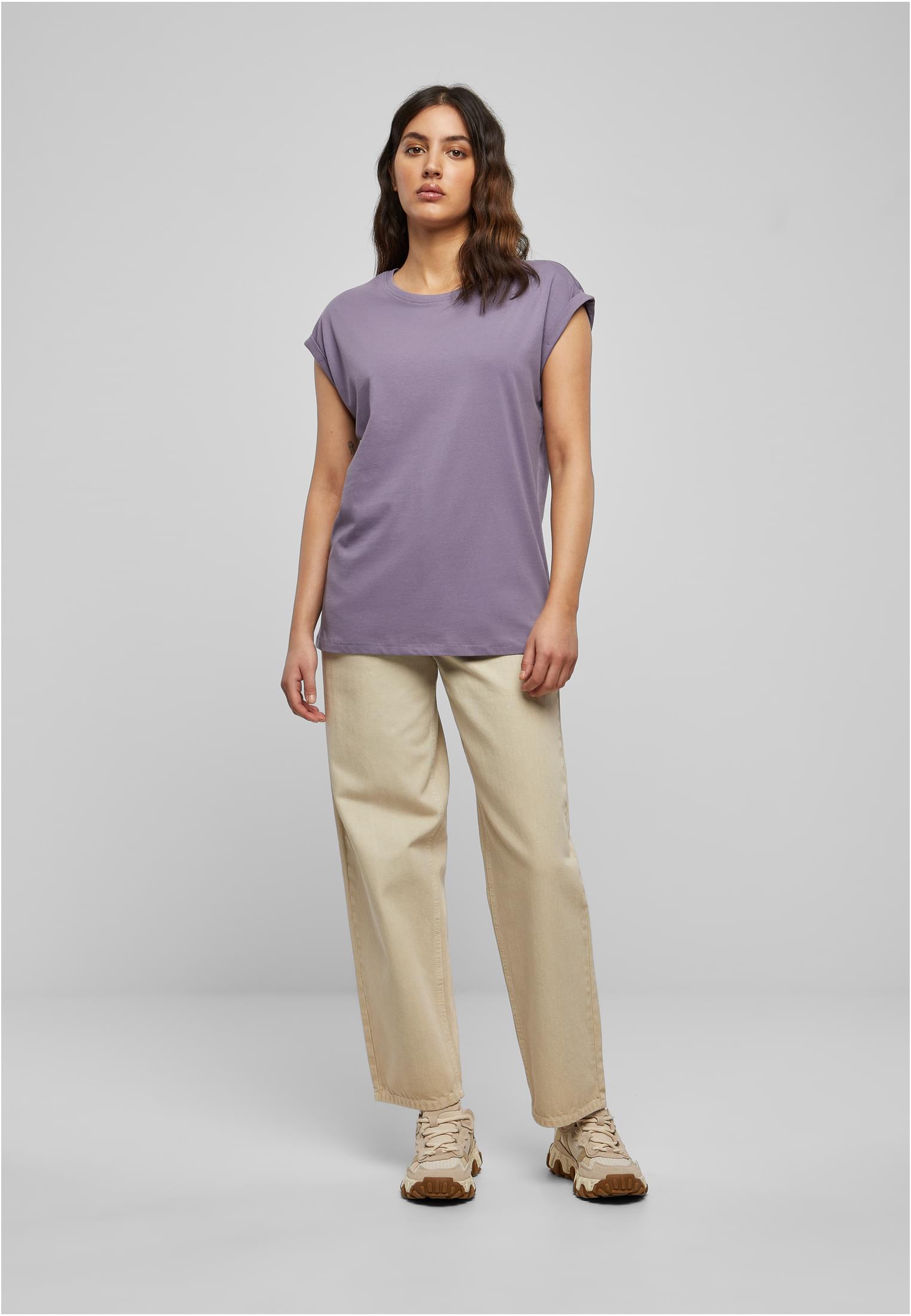 Kvinna i helfigur som poserar i en ljuslila Ladies Extended Shoulder Tee, beiga byxor och sneakers mot en grå bakgrund.