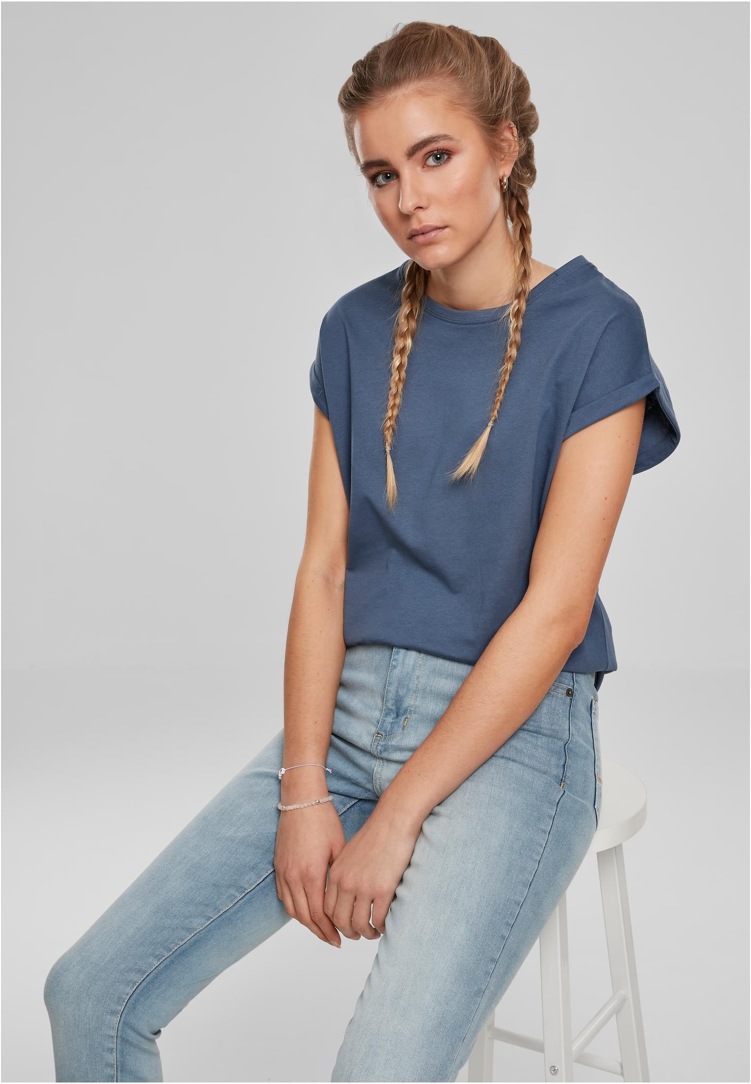 En kvinna med blonda flätor poserar sittandes på en pall, klädd i en blå Ladies Extended Shoulder Tee och ljusa jeans.