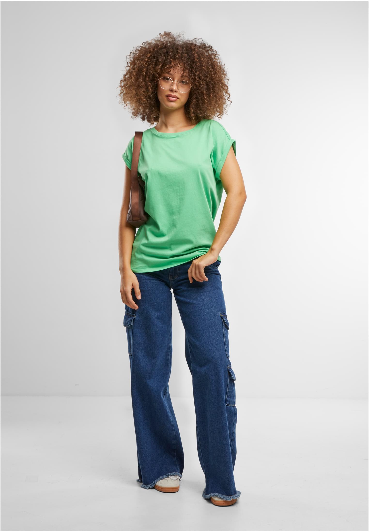 En kvinna med lockigt hår och glasögon poserar i en ljusgrön t-shirt med nedhasade axlar och mörkblå jeans med vida ben, mot en vit bakgrund.
